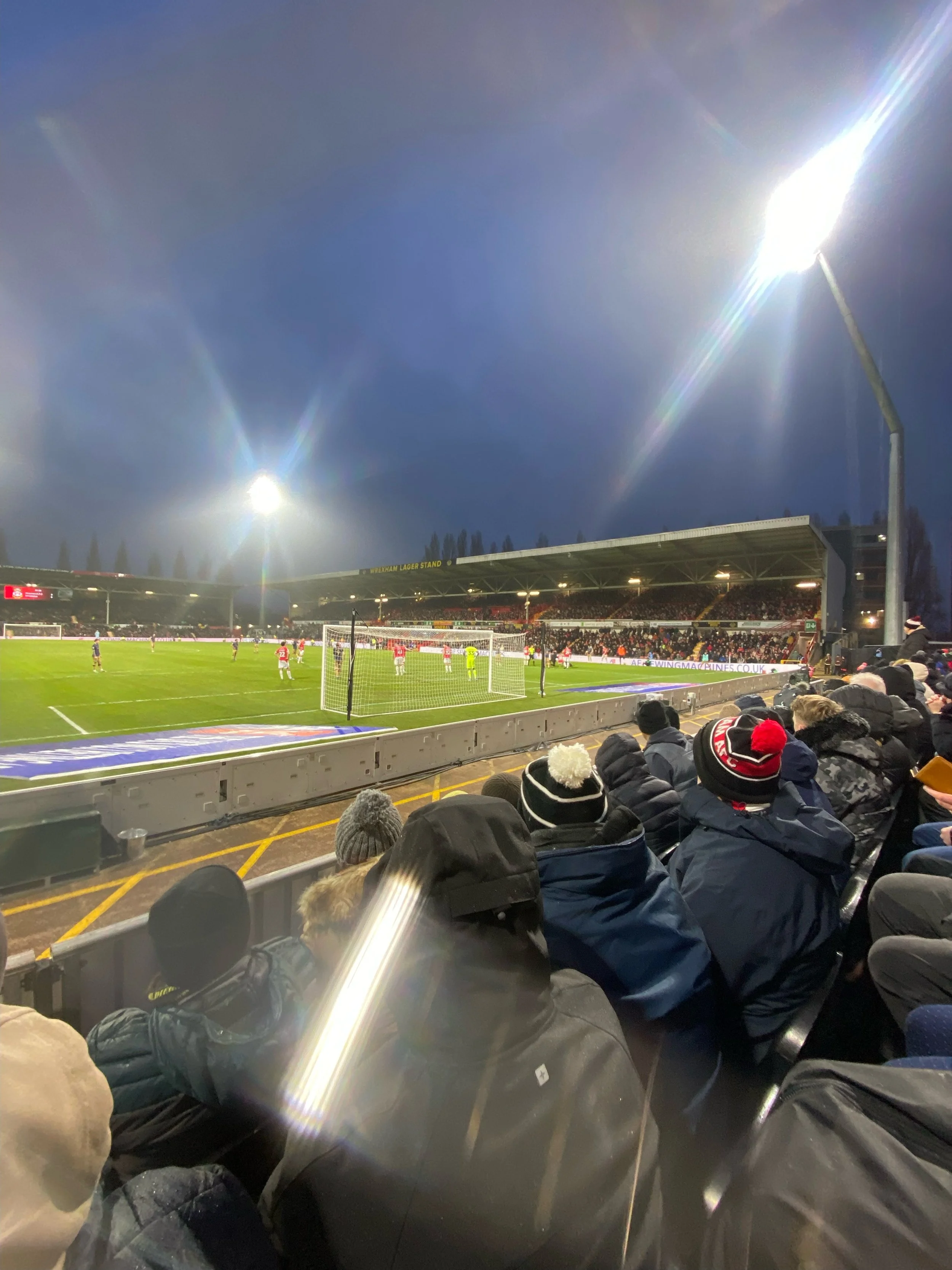 Crowd of spectators watching a soccer game at a stadium during dusk, with players on the field and stadium lights illuminating the scene.