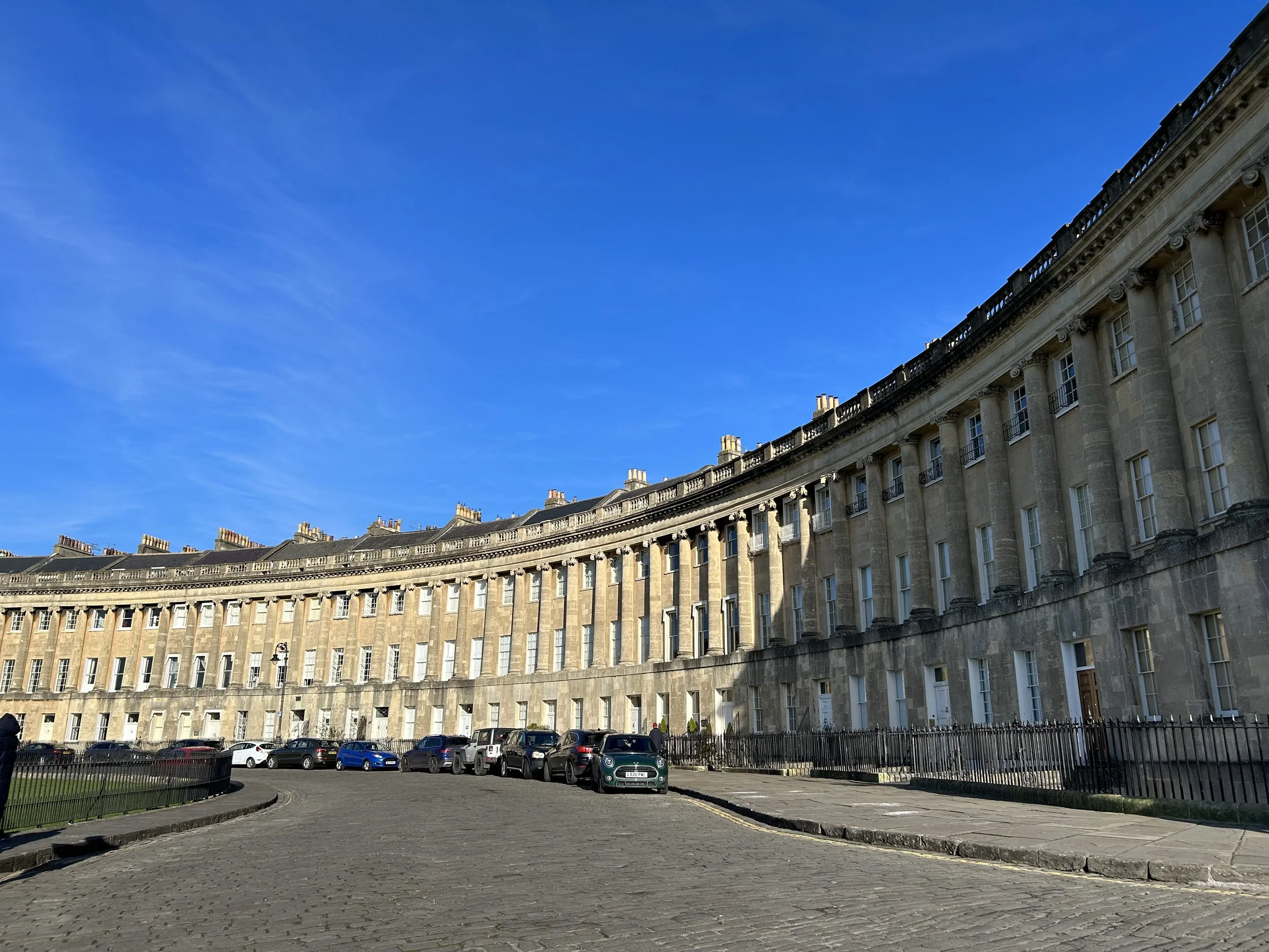 Curved row of historic terraced townhouses with beige stone facades and tall windows, under a bright blue sky, with parked cars along the cobblestone street.