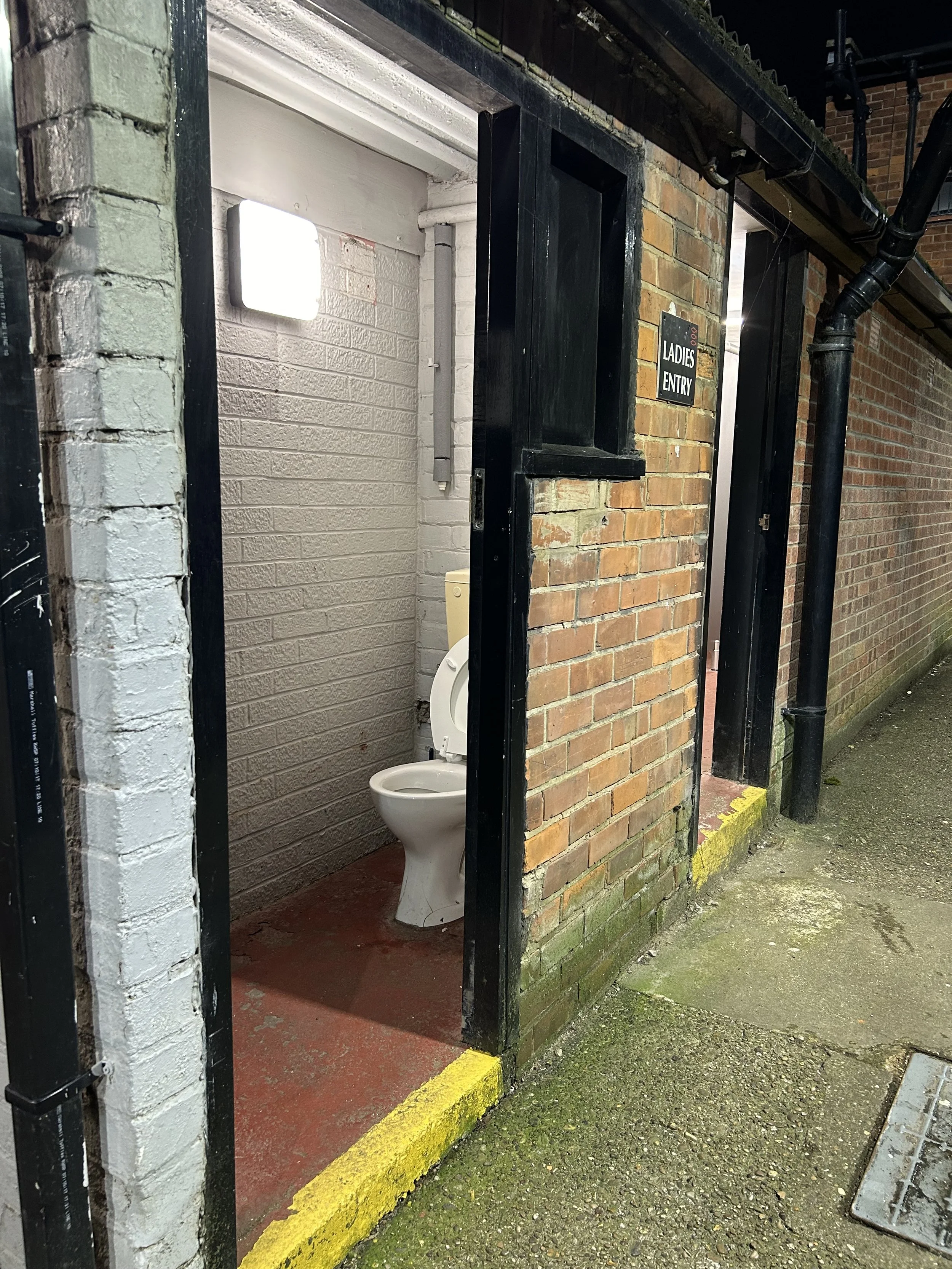 Outdoor women’s bathroom with an open door, a toilet inside, and a sign that reads "Ladies Entry" on the brick wall next to the door.