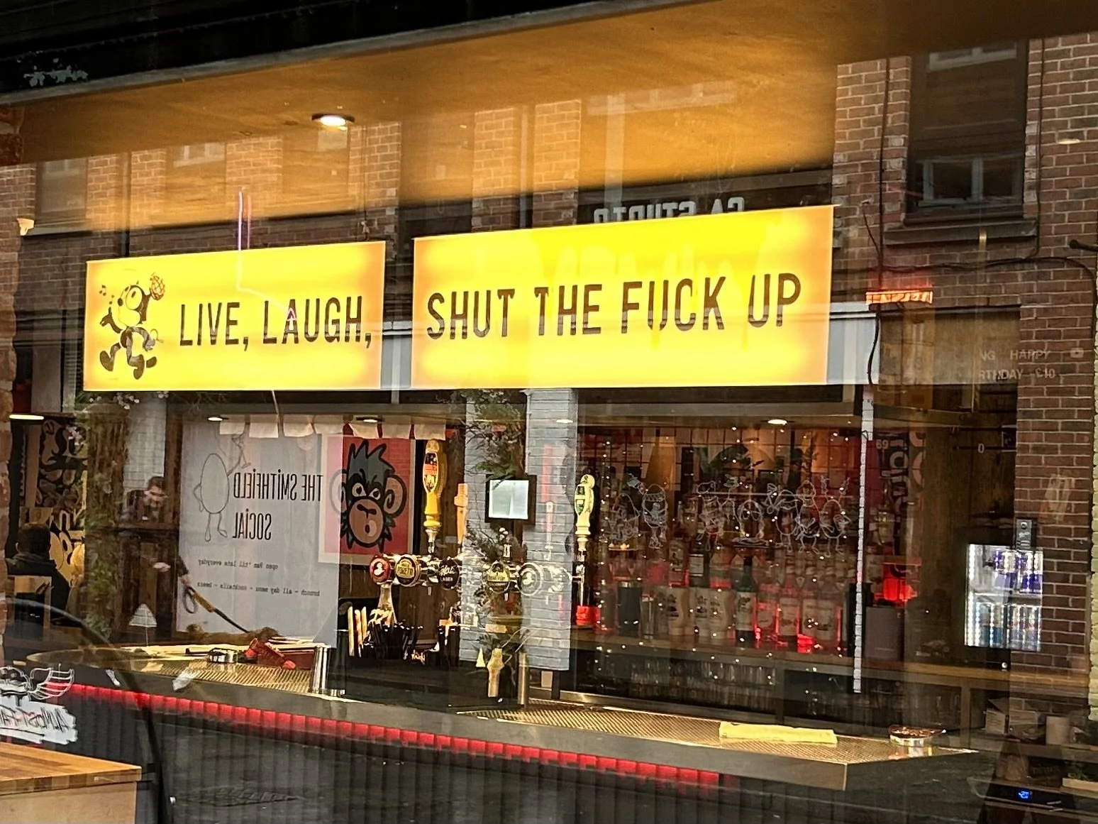 Reflection of a restaurant window with bright yellow signs reading 'Live, Laugh,' and 'Shut the F*** Up' in black text, with cartoon characters and restaurant interior visible inside.