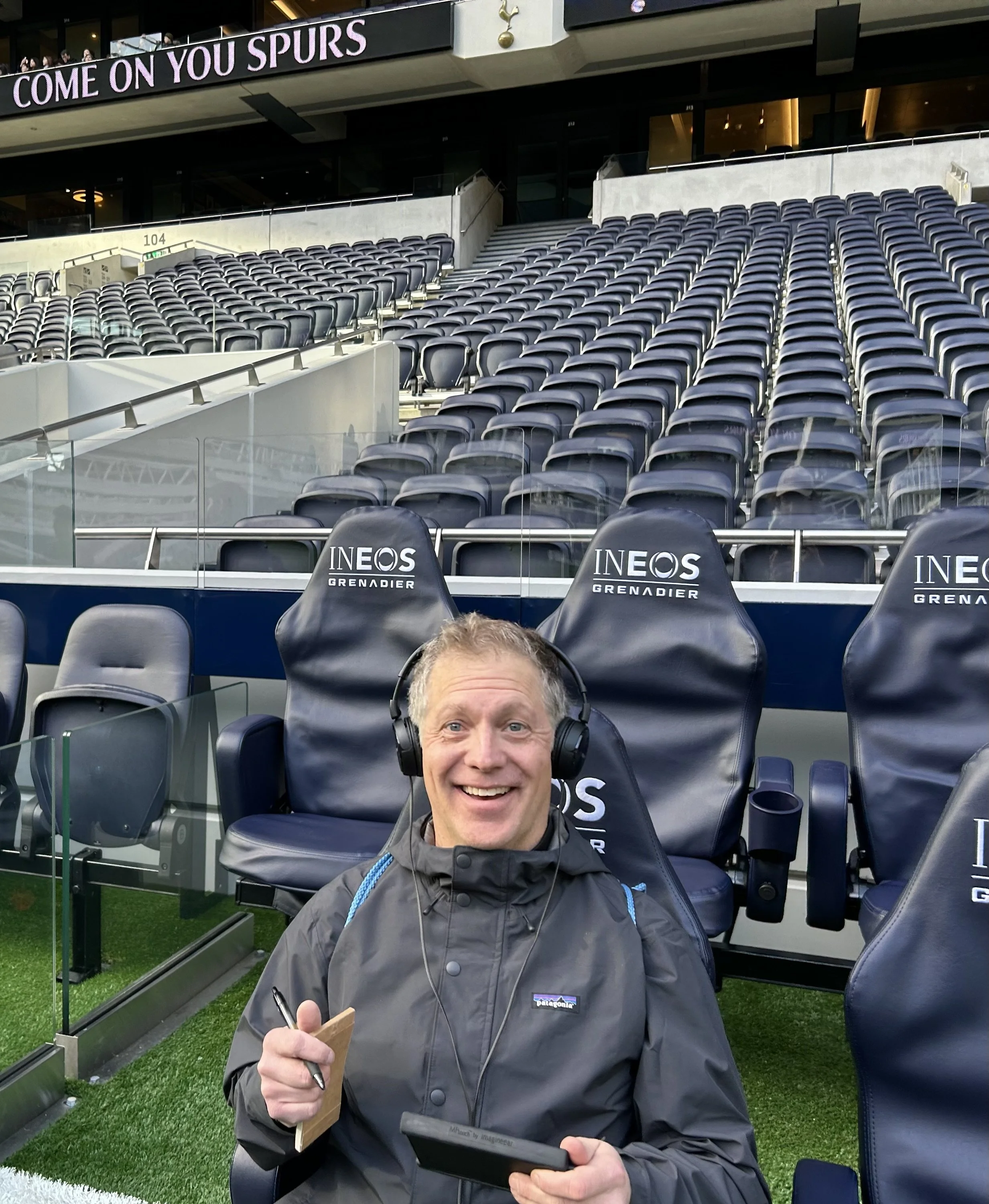 Author, Todd Smith smiling and wearing headphones, holding a notepad and pen, seated in front of empty seats at a football stadium, with a sign that reads 'COME ON YOU SPURS' visible in the background.