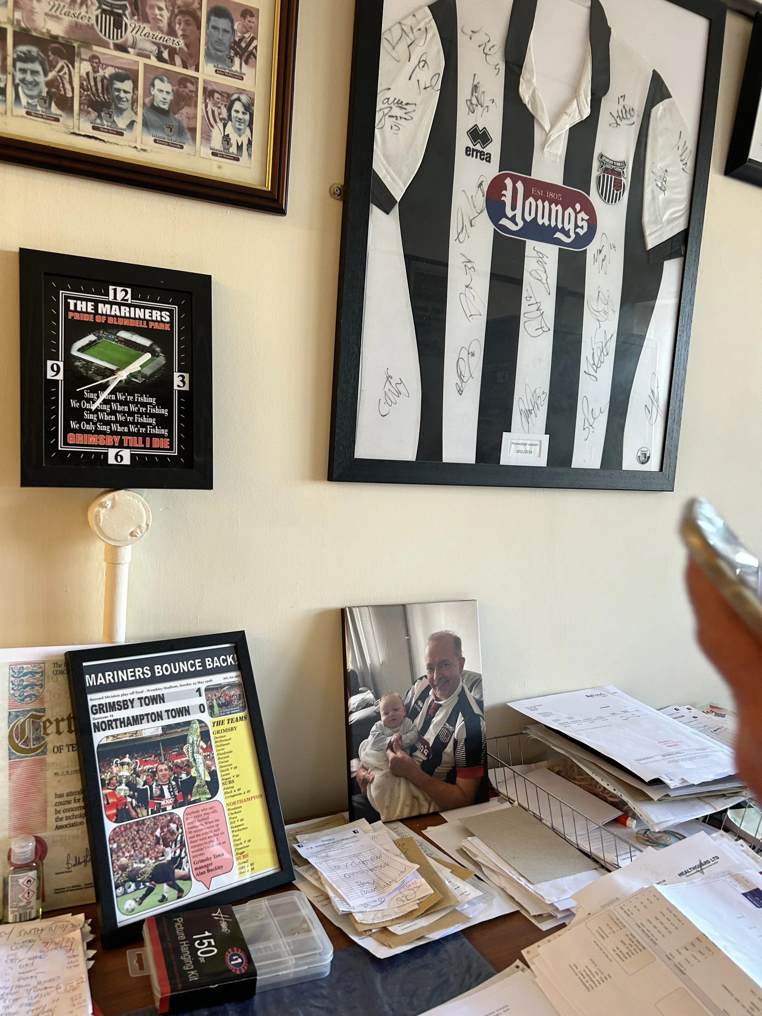A wall with framed items, including a framed signed football jersey, a signed photo of a person holding a baby, and a collage of mariner photos. A desk below the wall is cluttered with papers, a small plastic box, and a framed newspaper article.