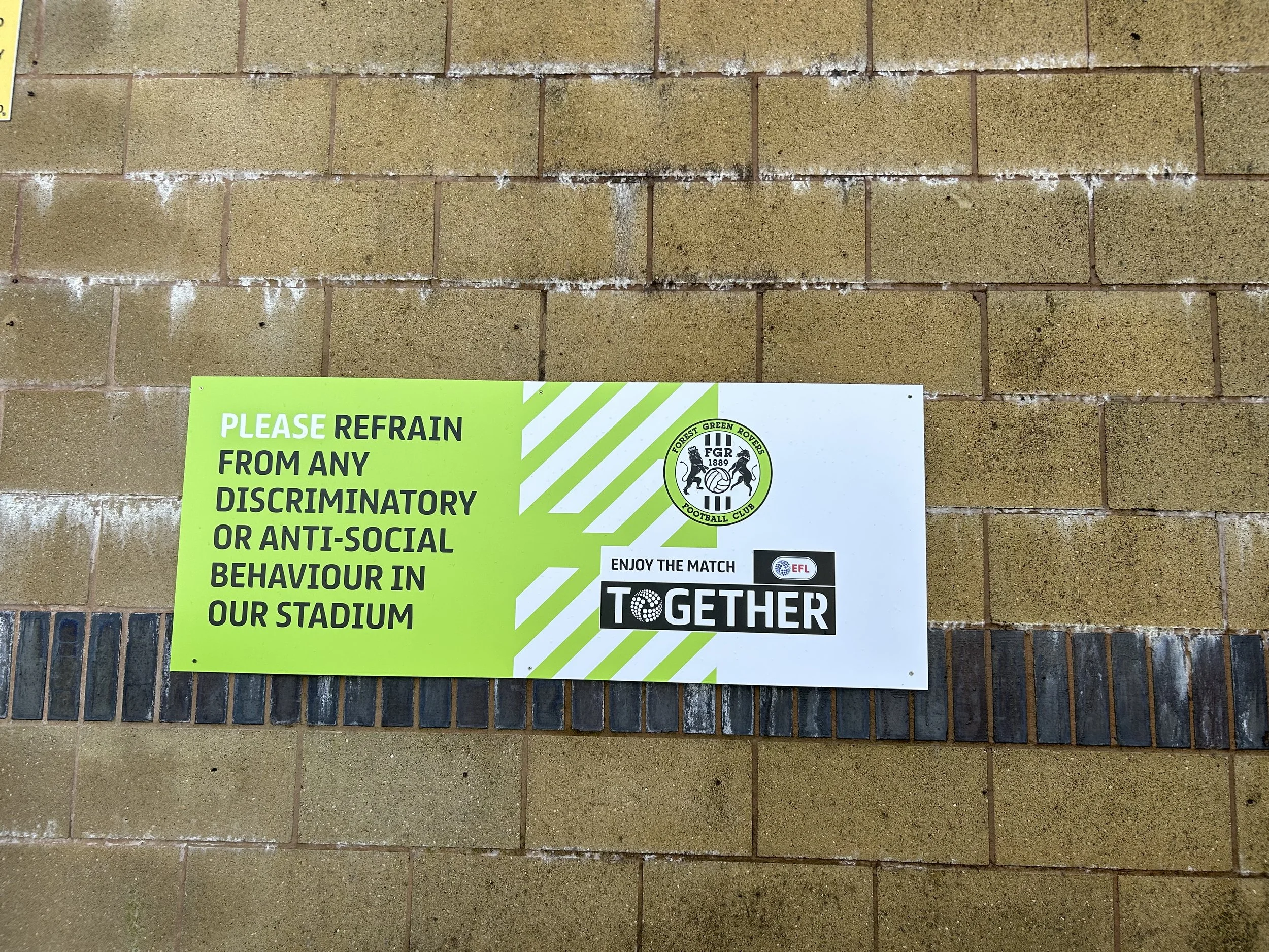 A sign on a brick wall requesting stadium visitors to refrain from any discriminatory or anti-social behavior, with logos of Forest Green Rovers Football Club and EFL, and a message to enjoy the match together.