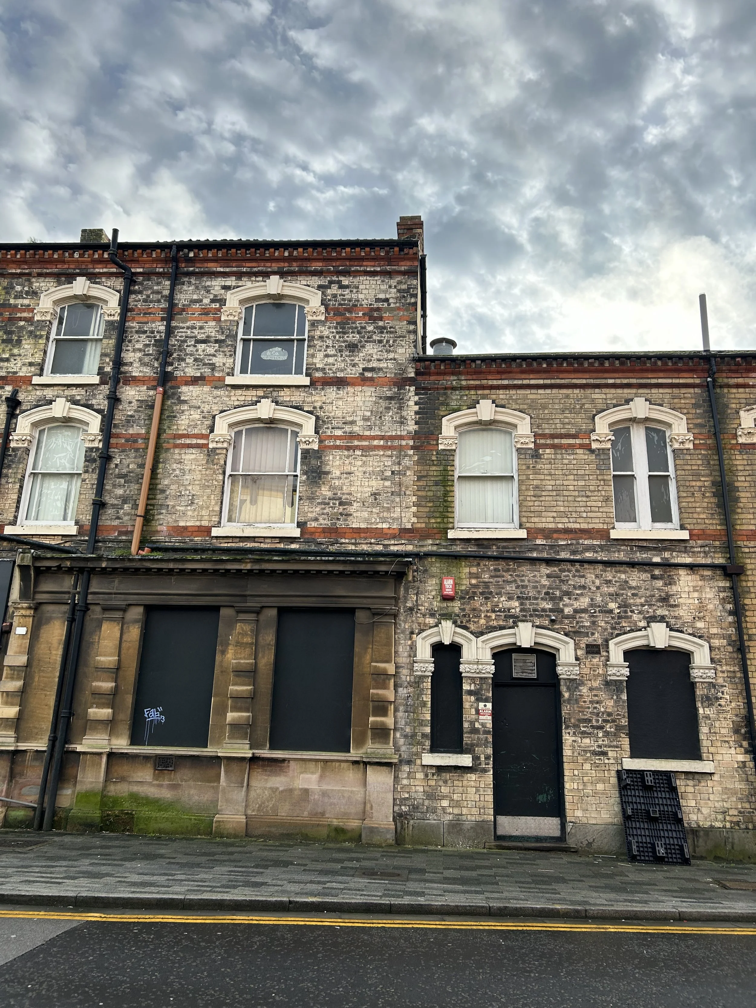 Three-story brick building with boarded windows and arched window frames, showing signs of wear and moss, under a cloudy sky.