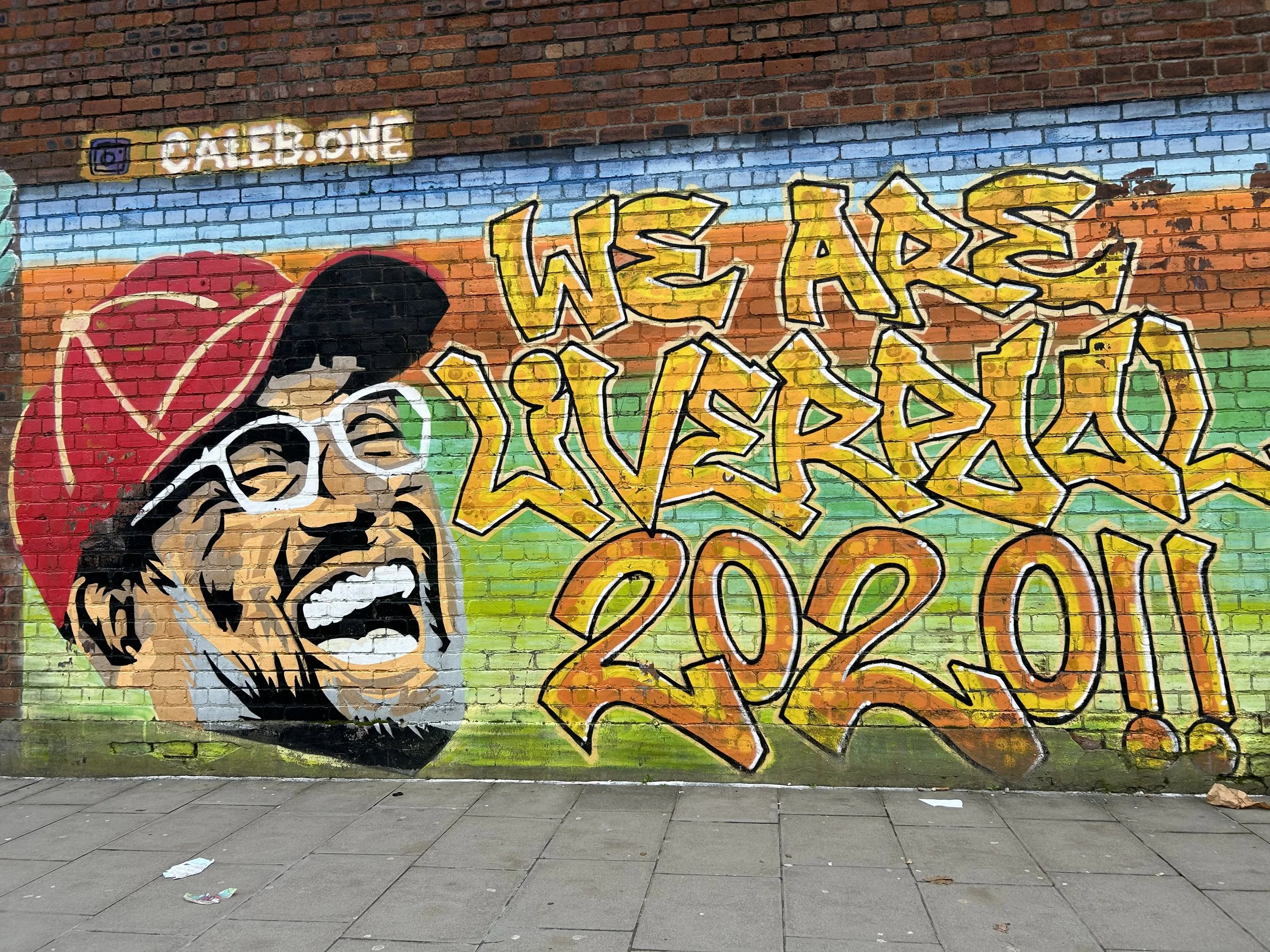 Graffiti art on a brick wall with a shouting man's face wearing glasses and a red hat. Text in yellow and orange graffiti reads "WE ARE LIVERMORE 2021" and a small Instagram handle "QALB.ONE" is at the top.