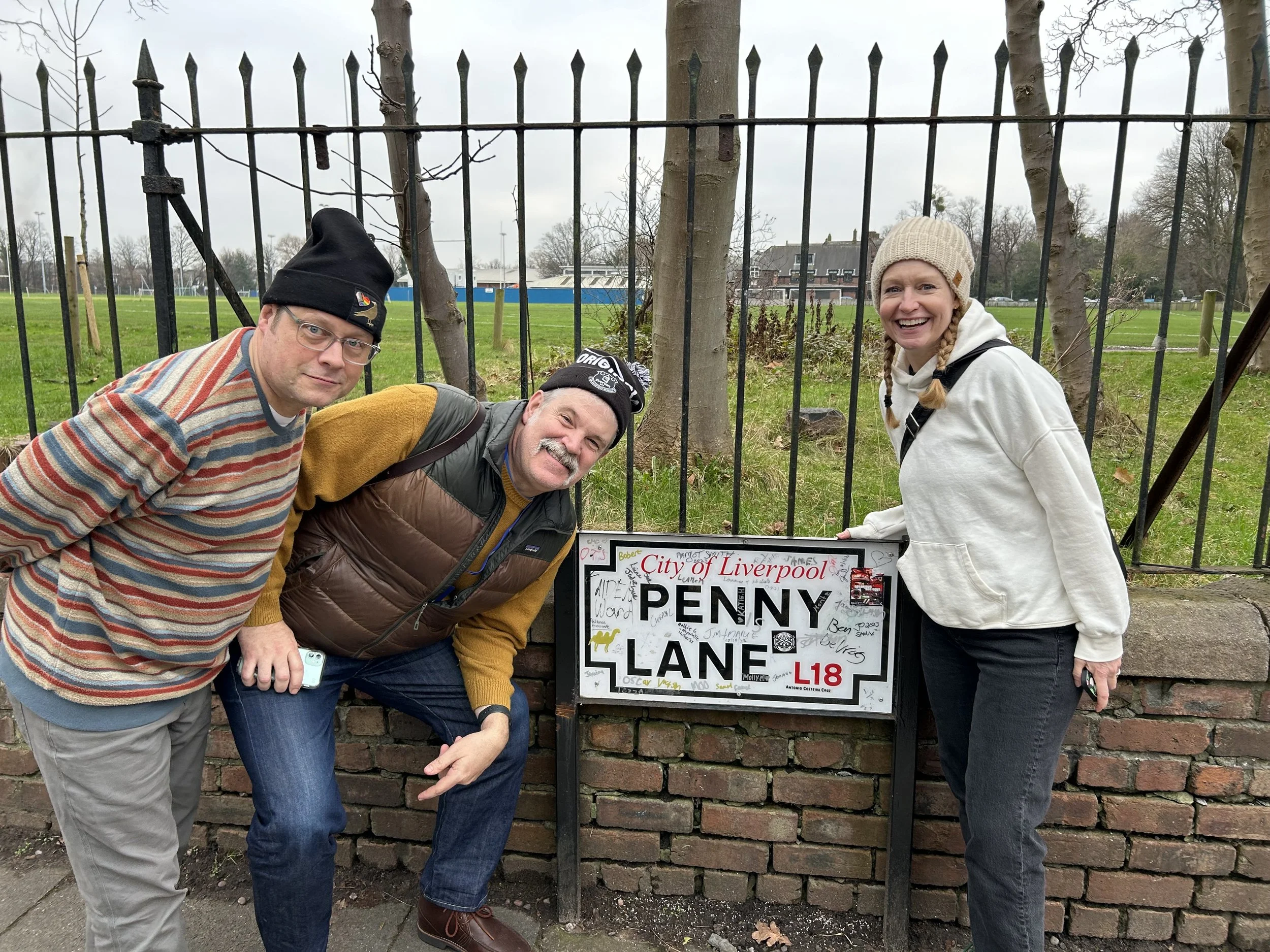 Todd Smith and wife standing outside next to a sign that reads 'City of Liverpool Penny Lane L18'. They are posing for the photo, with a fence and trees in the background.