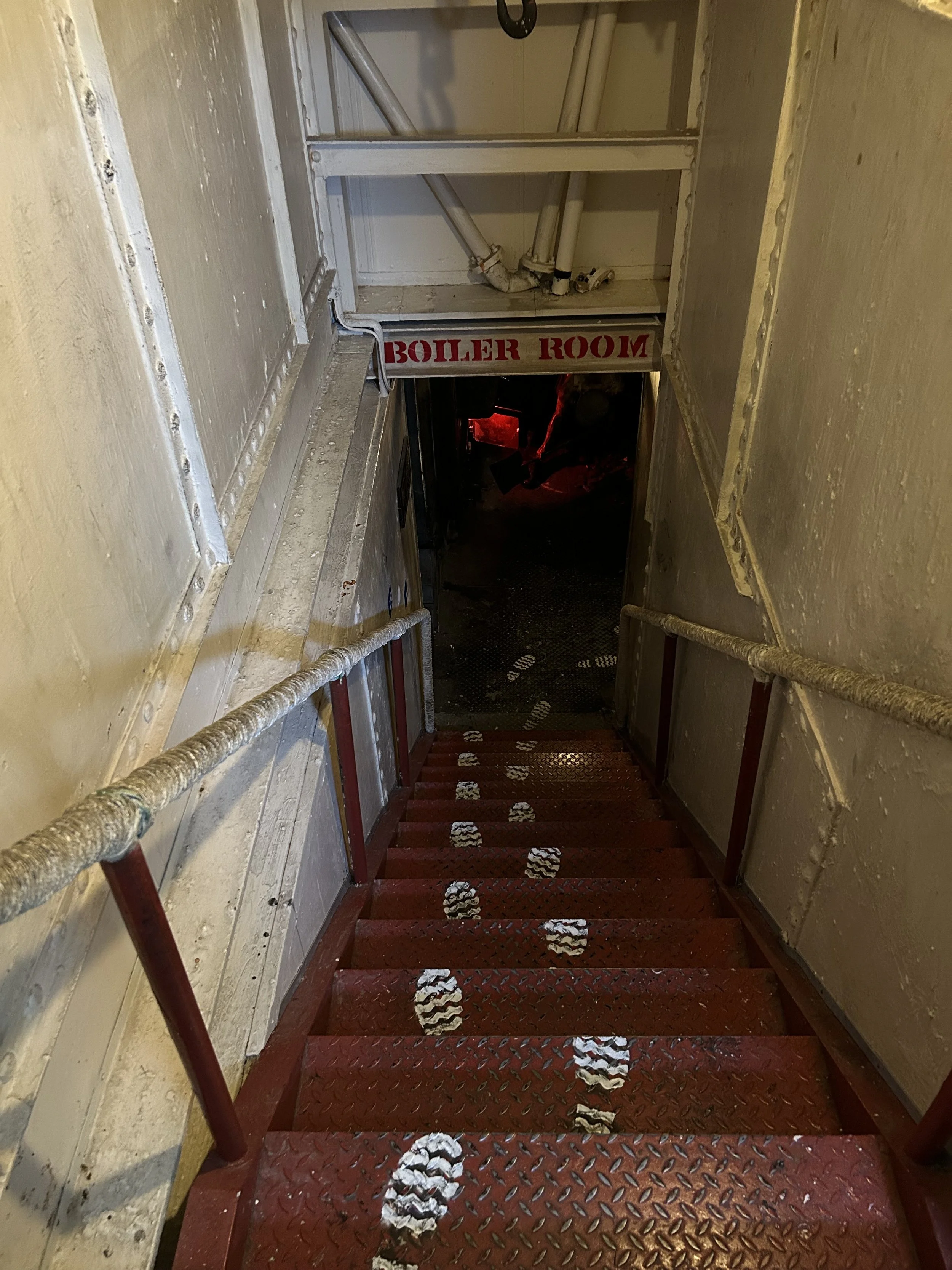 Downward view of a red metal staircase with white footprints painted on steps, leading to a door labeled 'Boiler Room' in red lettering. The staircase is enclosed by beige walls and has beige handrails on both sides.