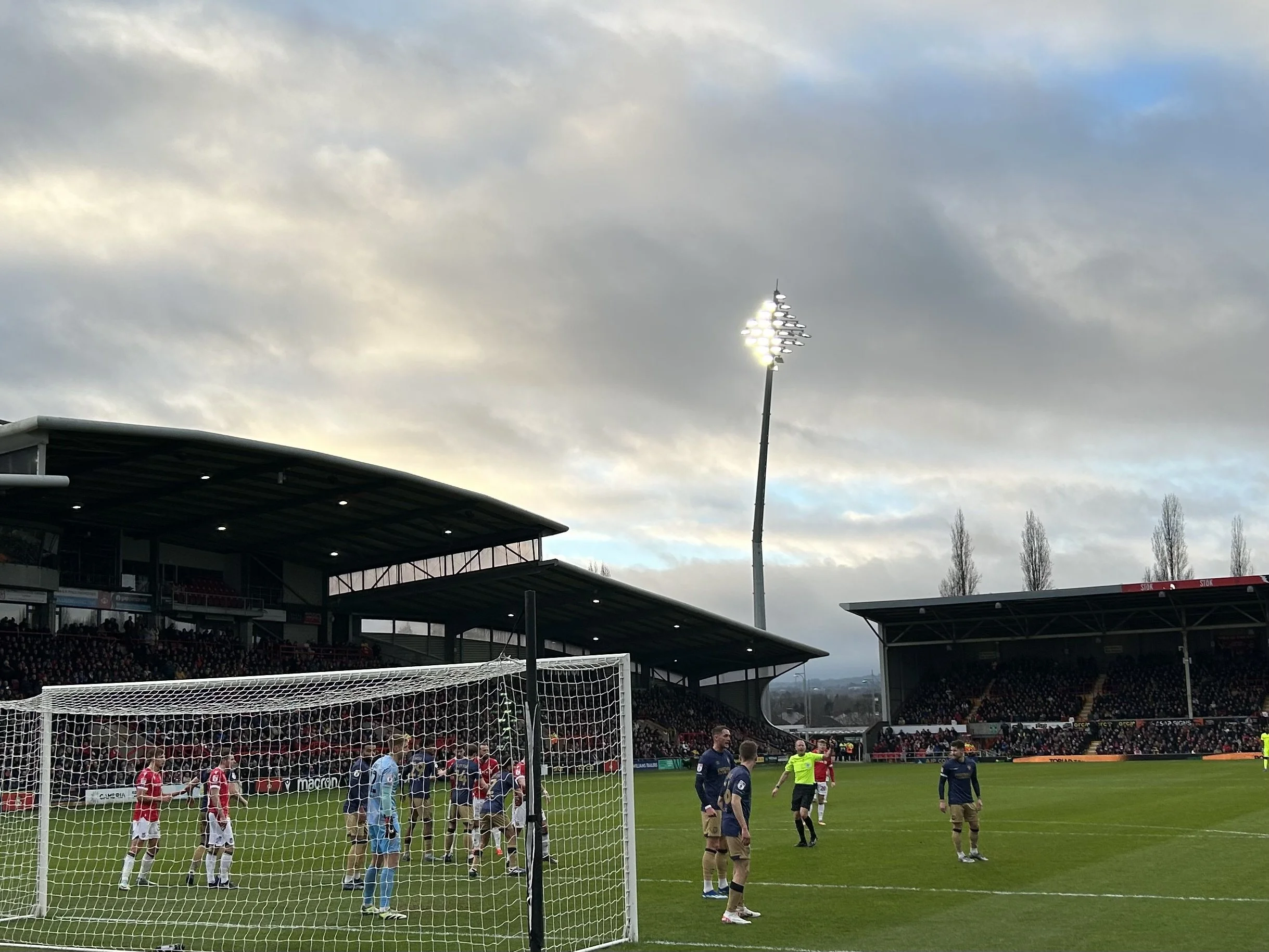 Soccer match in progress at a stadium during daytime, with players on the field, spectators in the stands, and stadium lights illuminating the scene under a cloudy sky.