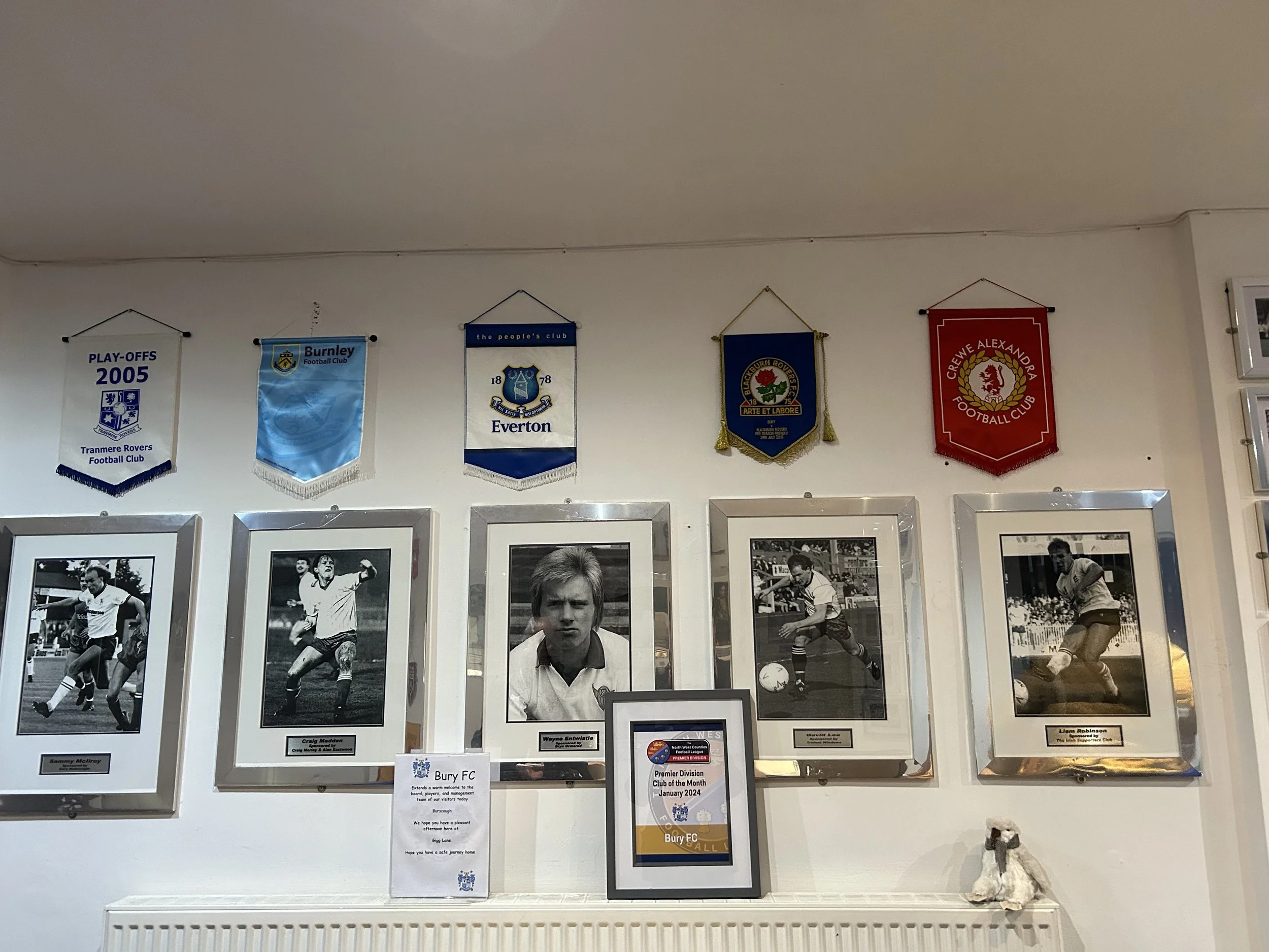 Wall display of framed black-and-white photographs of football players, with a series of colorful football club banners hanging above, including banners for Tranmere Rovers, Burnley, Everton, Leicester City, and Crewe Alexandra.