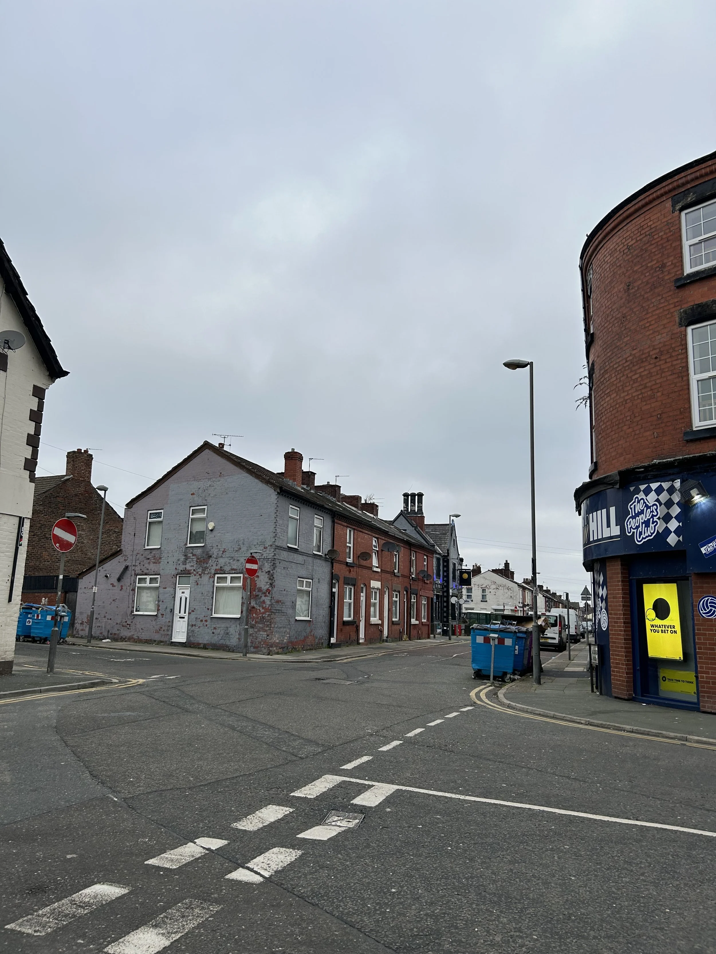 This is an urban street scene with a corner building on the right and a row of houses on the left. The sky is overcast, and there are street signs, parked cars, and trash bins visible.