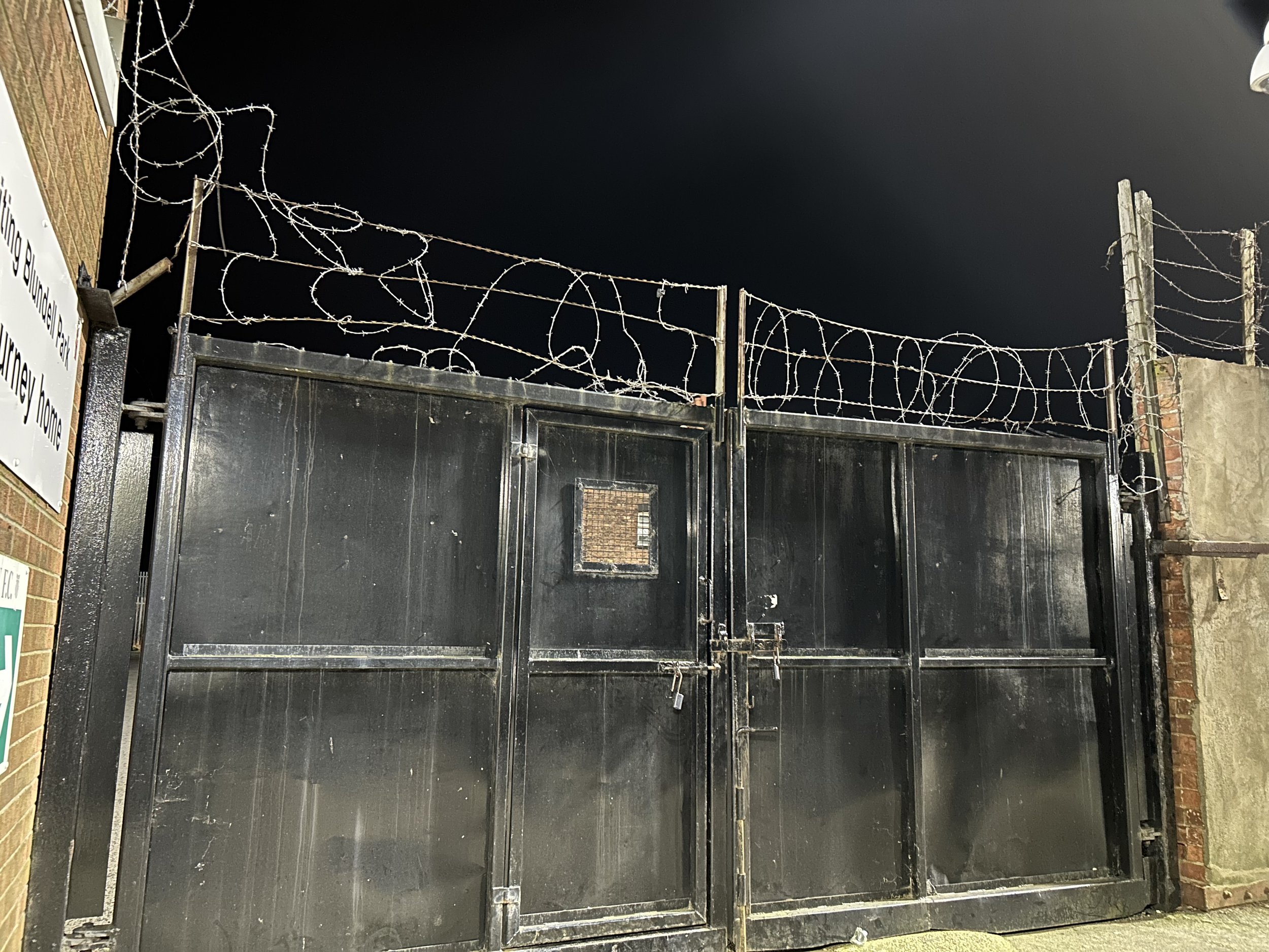 Black metal gate topped with barbed wire, situated between brick and concrete walls at night.