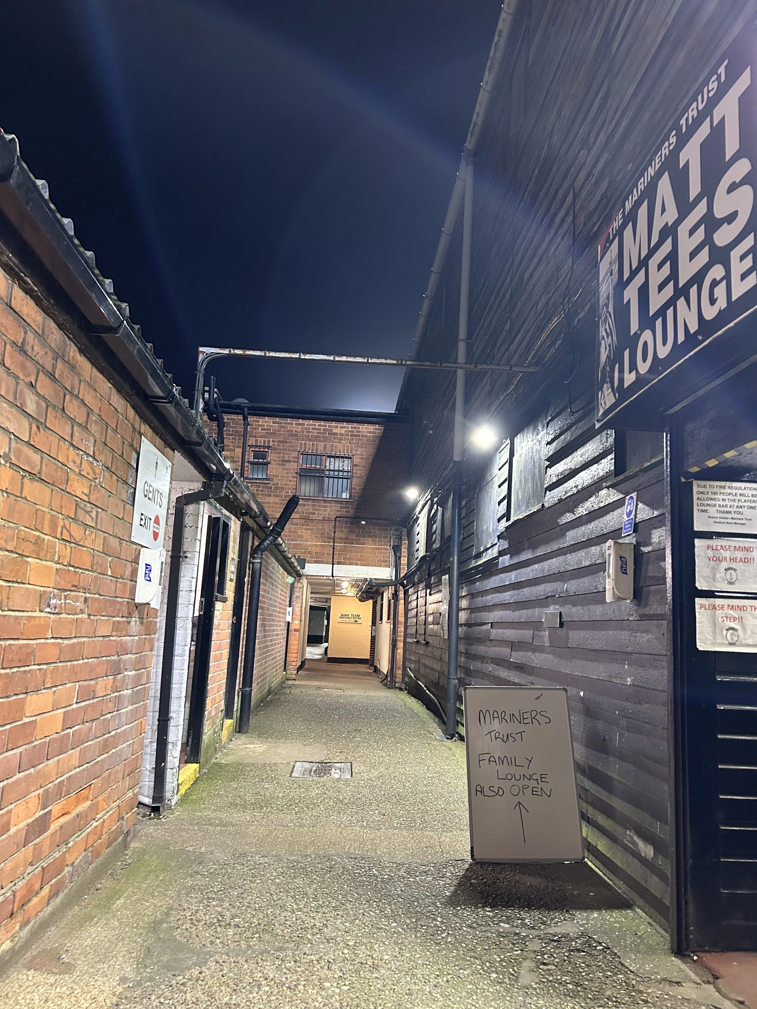 Night view of an alleyway with brick and wooden walls, illuminated by overhead lights. A sign on the ground indicates the Mariners Trust Family Lounge is open. There are signs regarding adult and children’s sections and a notice about access restrict