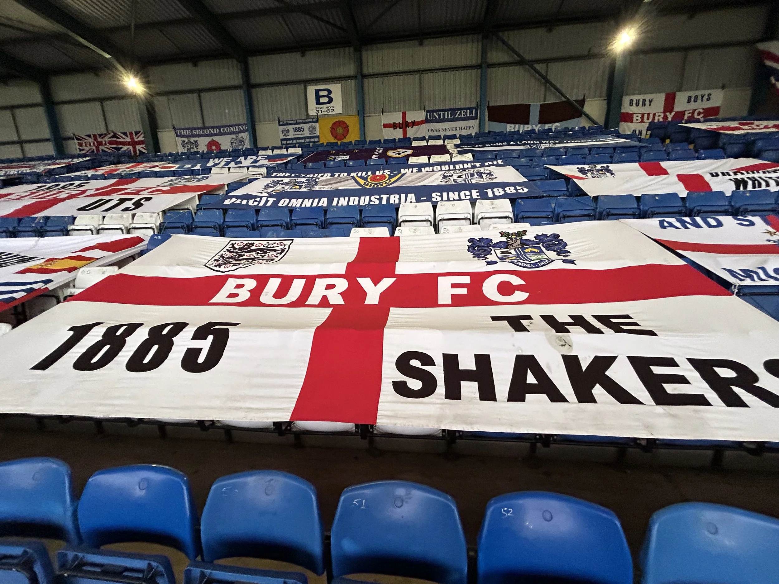 A collection of Bury FC banners and flags draped over stadium seats, including one prominent large flag with the club's name, emblem, and the year 1885.