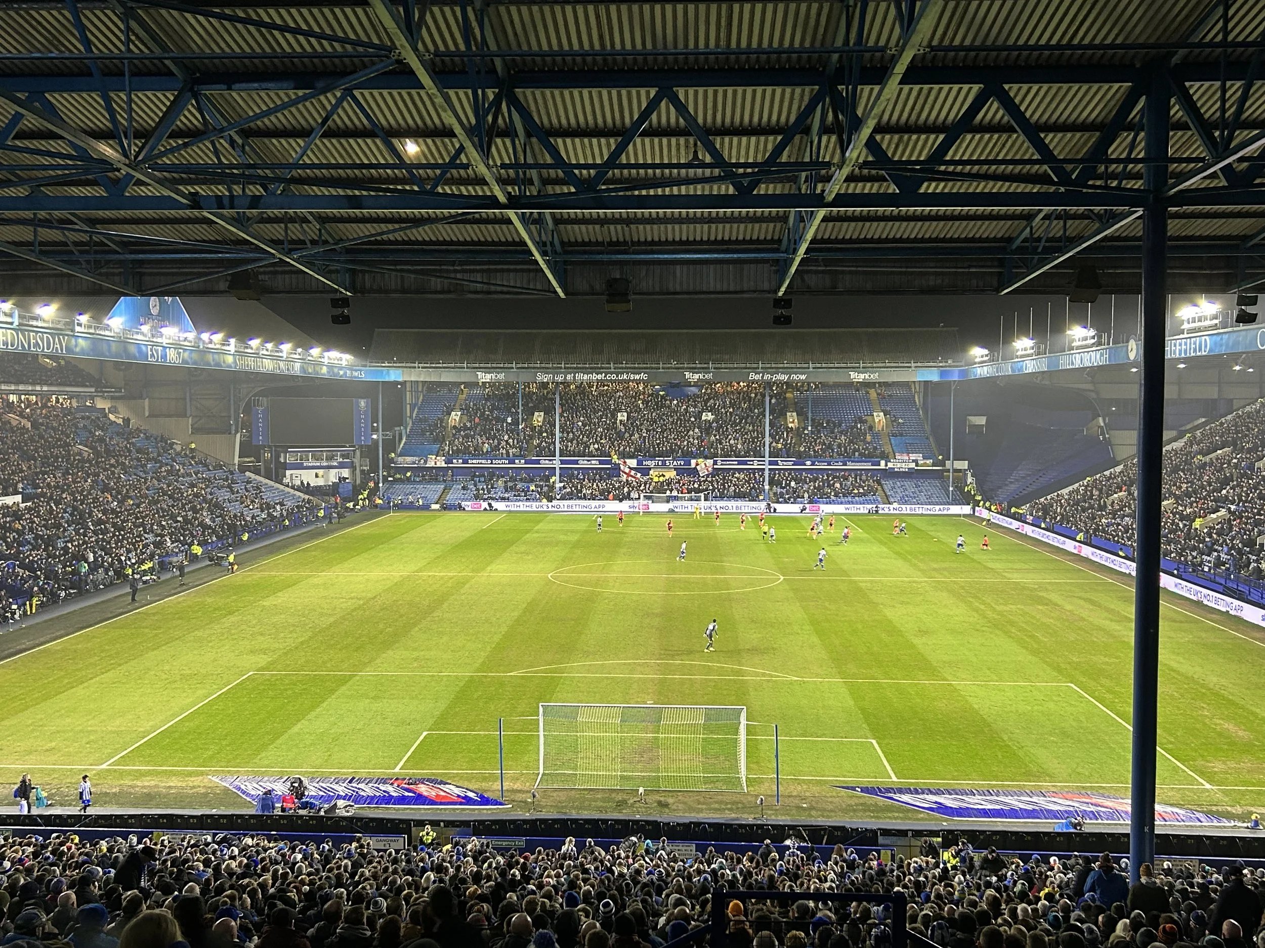 A nighttime view of a soccer stadium filled with spectators, with players on the field. The stadium has a large roof structure and advertising boards around the perimeter.