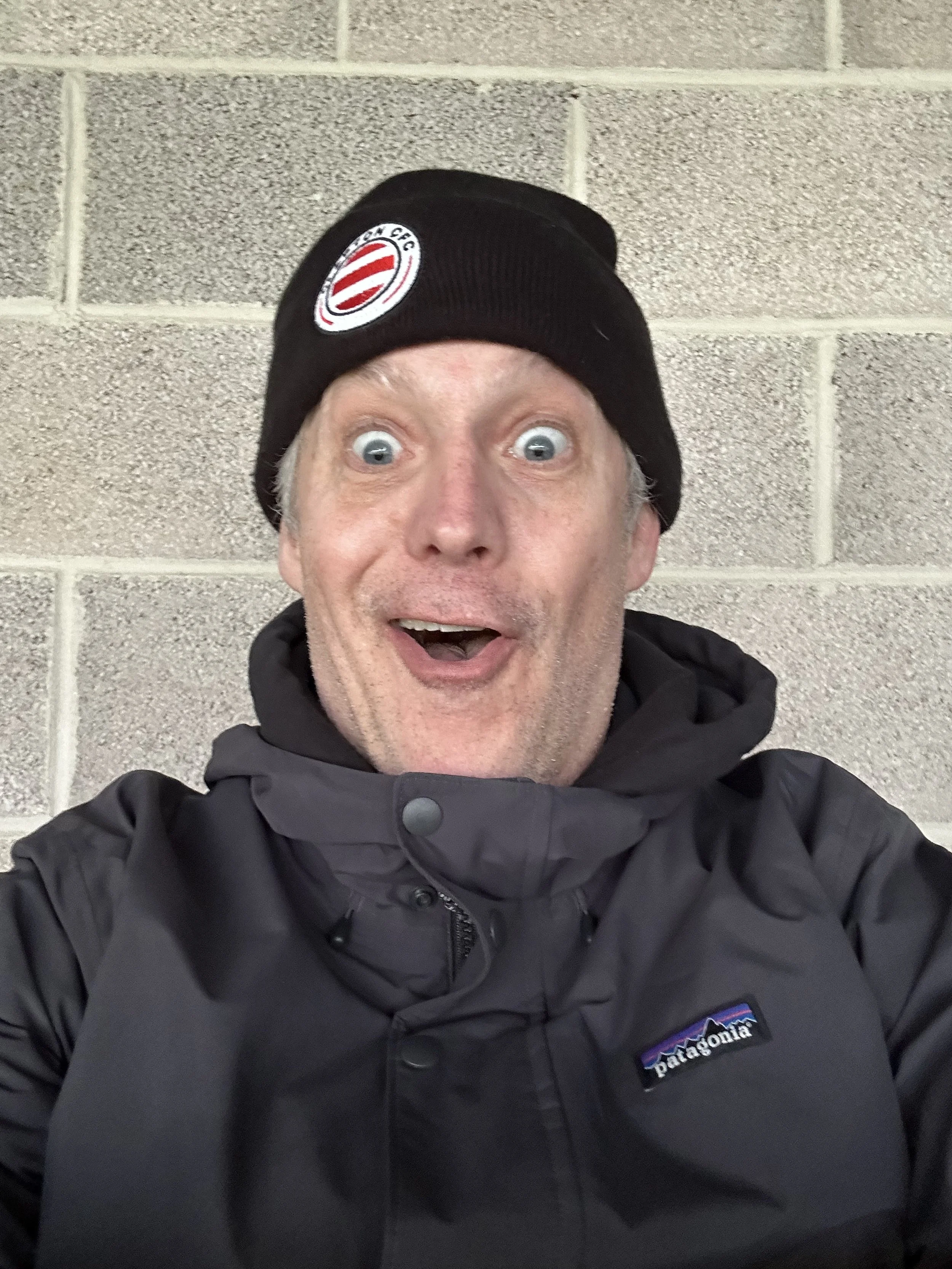 A man wearing a black beanie with a sports team logo, a black Patagonia jacket, and making an excited face, standing against a gray cinder block wall.