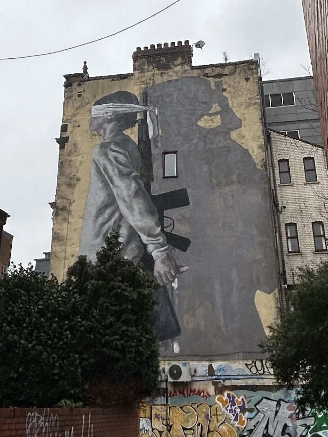 Large mural on building wall depicting two Black women in profile, one with a bandana over her eyes holding a rifle, the other with a hand near her face, painted in grayscale with yellow background details.