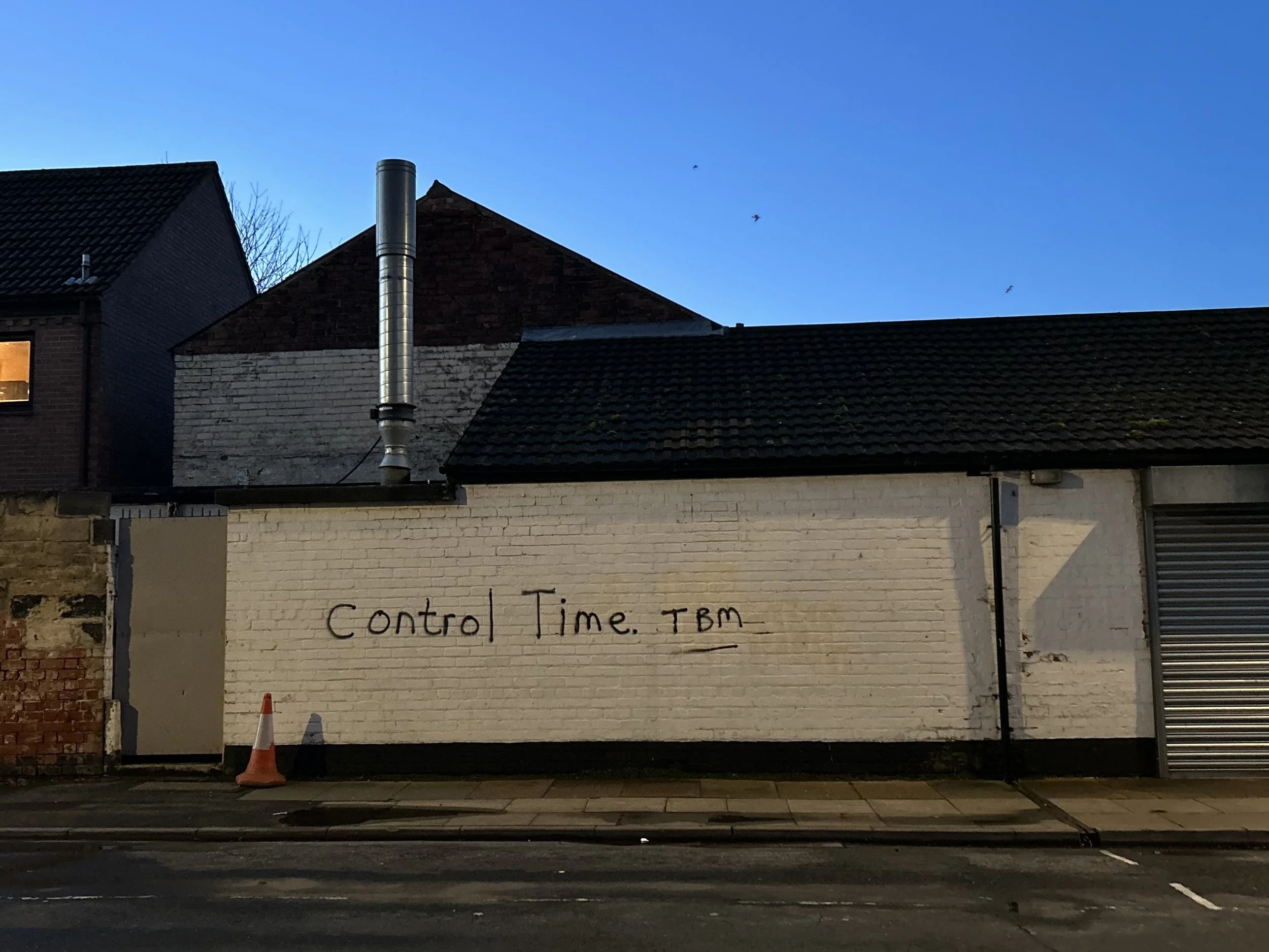 A brick building with graffiti reading 'Control Time. TBM' on a white-painted section of the wall, a traffic cone in front, in a neighborhood at dusk, with a clear sky and a couple of birds flying.