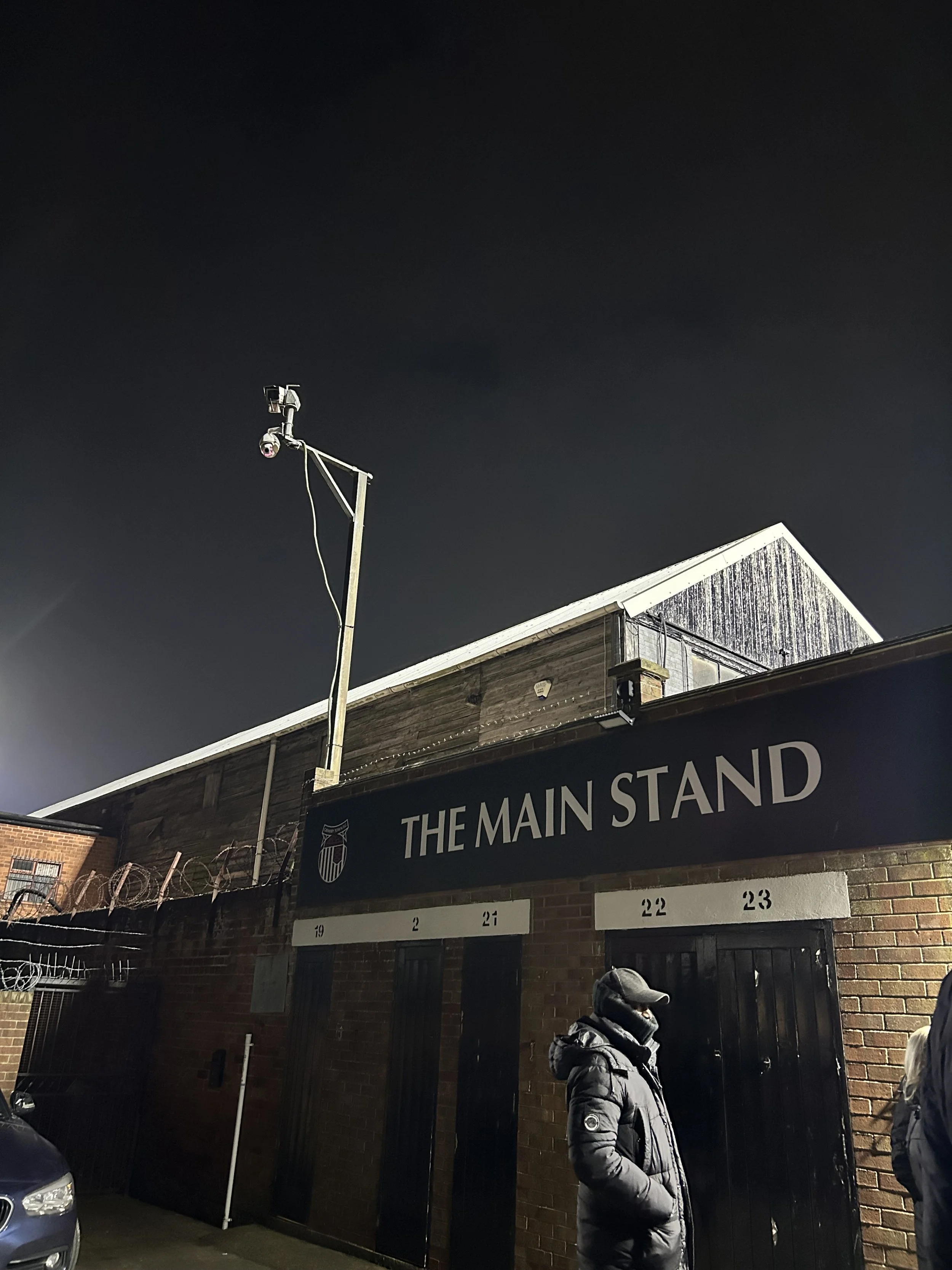 A building with a sign reading 'The Main Stand' and a person wearing a puffer jacket and cap standing outside at night.