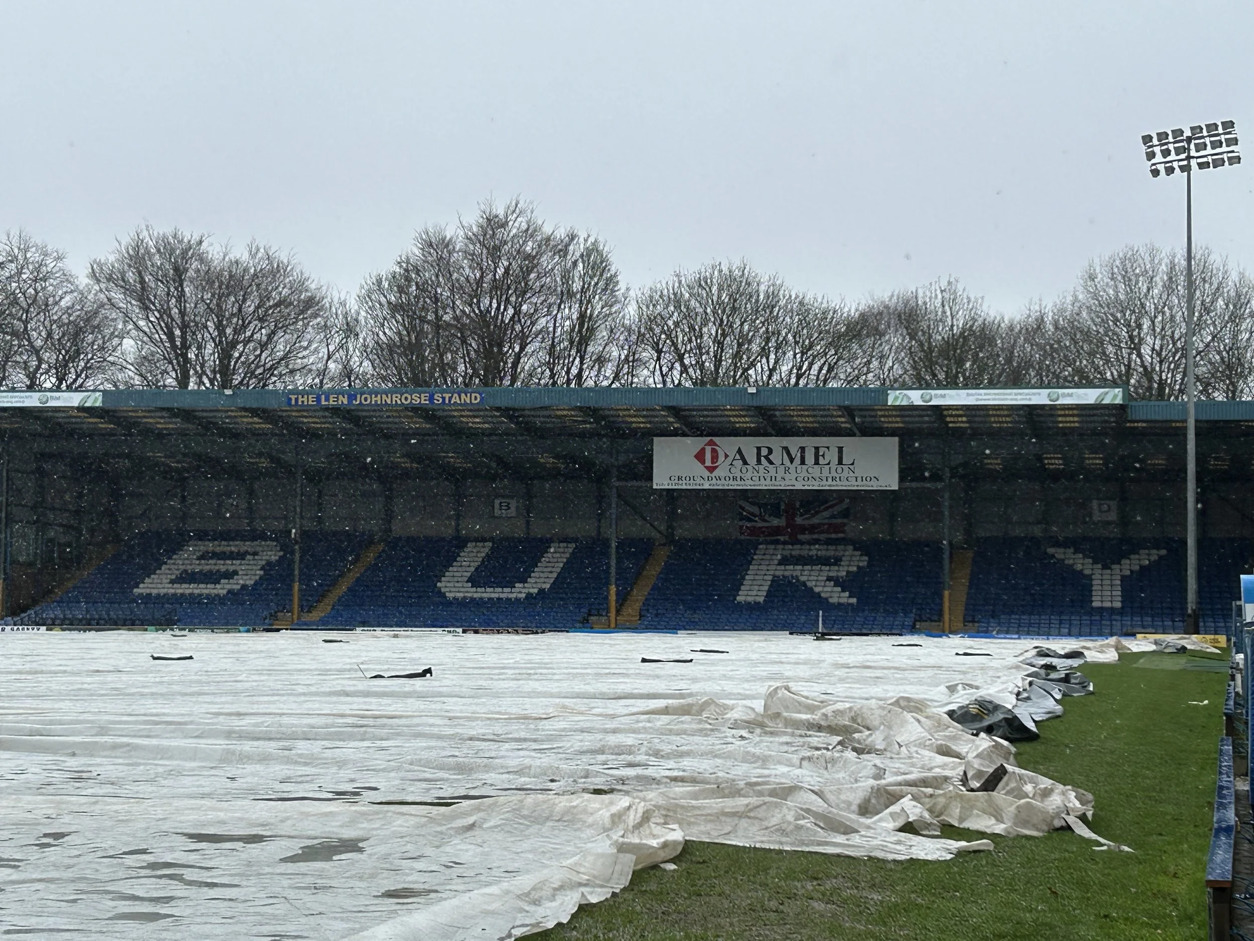 A football stadium with empty seats, a tarp covering the field, and trees in the background, under a gray sky.