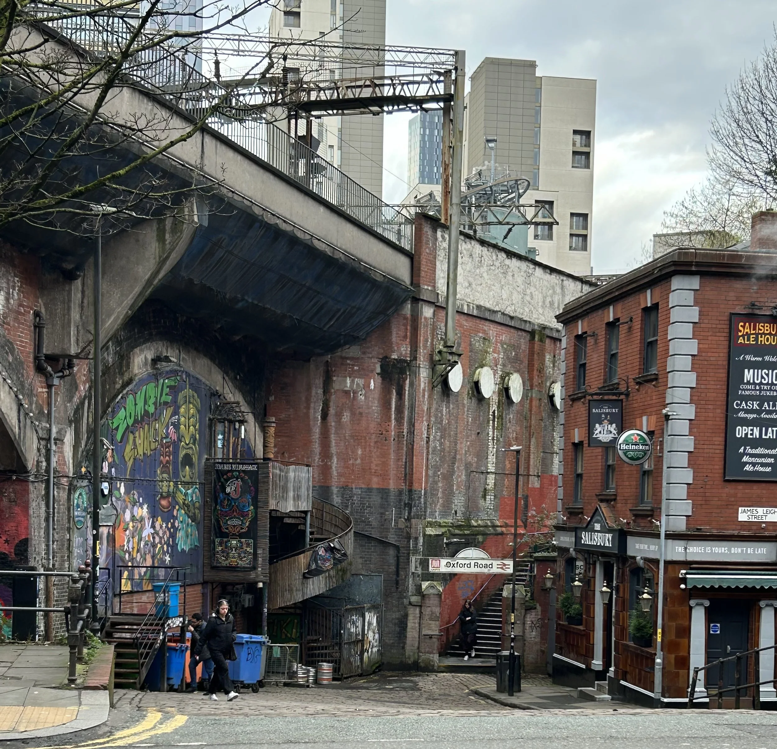 Street view of a brick building with a sign for Salisbury Ale House, next to a mural-covered underpass, stairs, and pedestrians in an urban area.