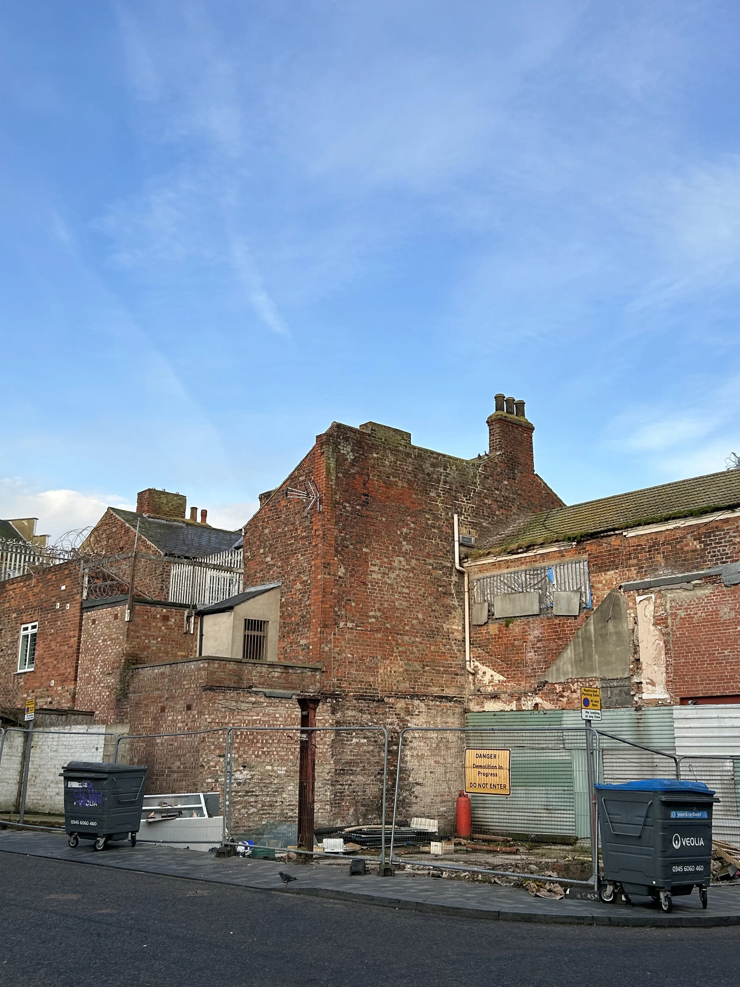 Old brick building under construction or demolition, surrounded by metal fencing with warning signs, trash bins, and construction debris, against a blue sky.