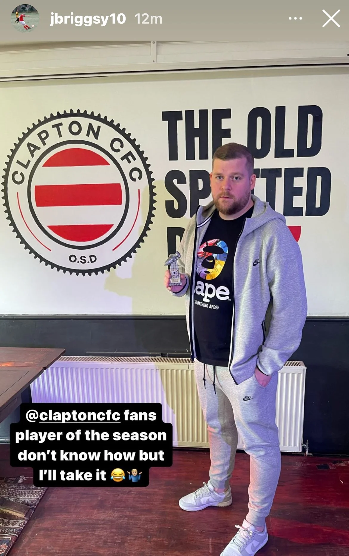 Man in gray hoodie and sweatpants holding a trophy, standing in front of an 80s-themed wall with the Clapton CFC logo and wording 'The Old Spected'.