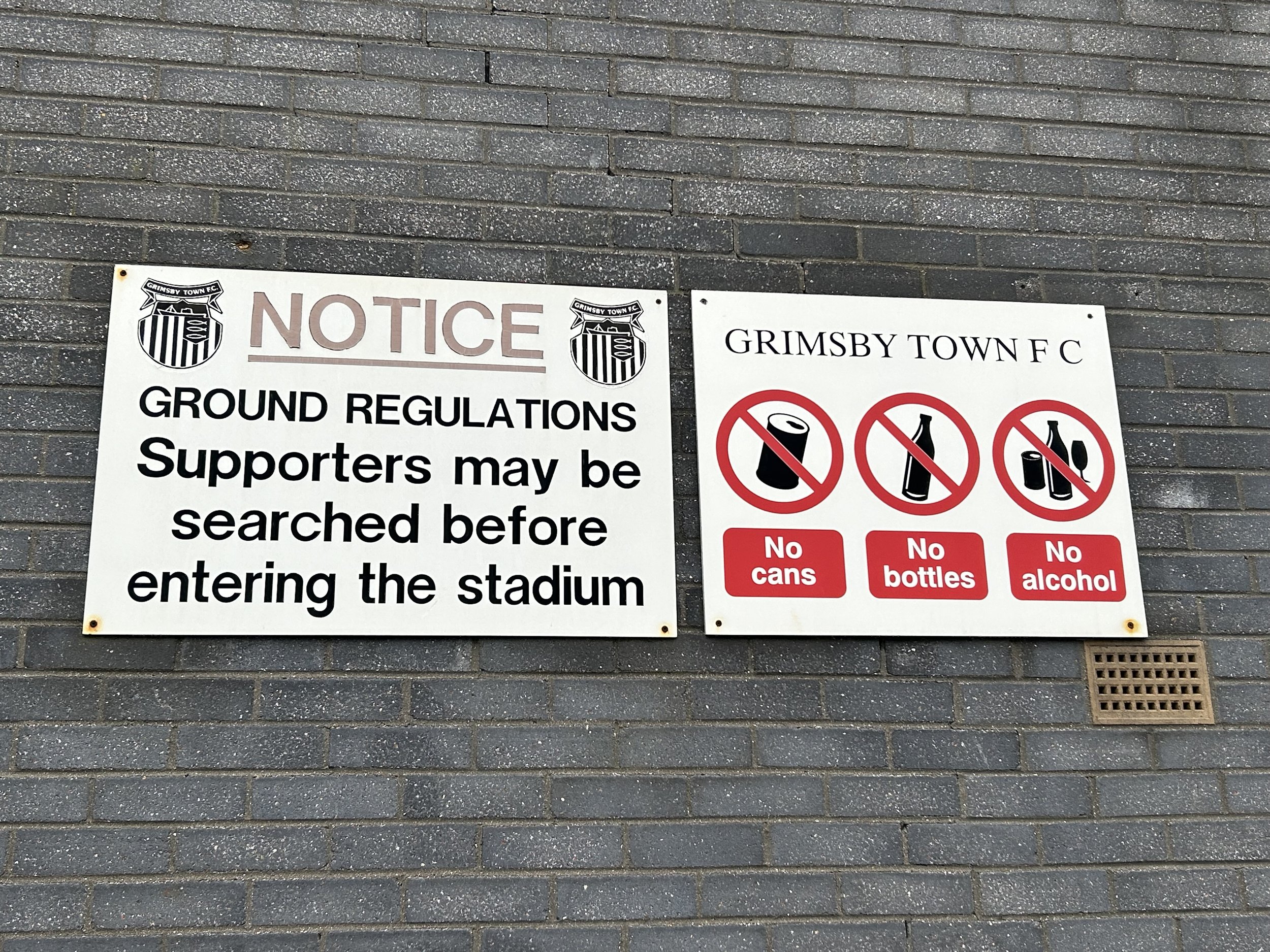 Sign on a brick wall with notices: one notice says 'NOTICE GROUND REGULATIONS Supporters may be searched before entering the stadium'. The other notice says 'GRIMSBY TOWN F C' with icons of a can, bottle, and alcohol crossed out, and labels 'No cans,