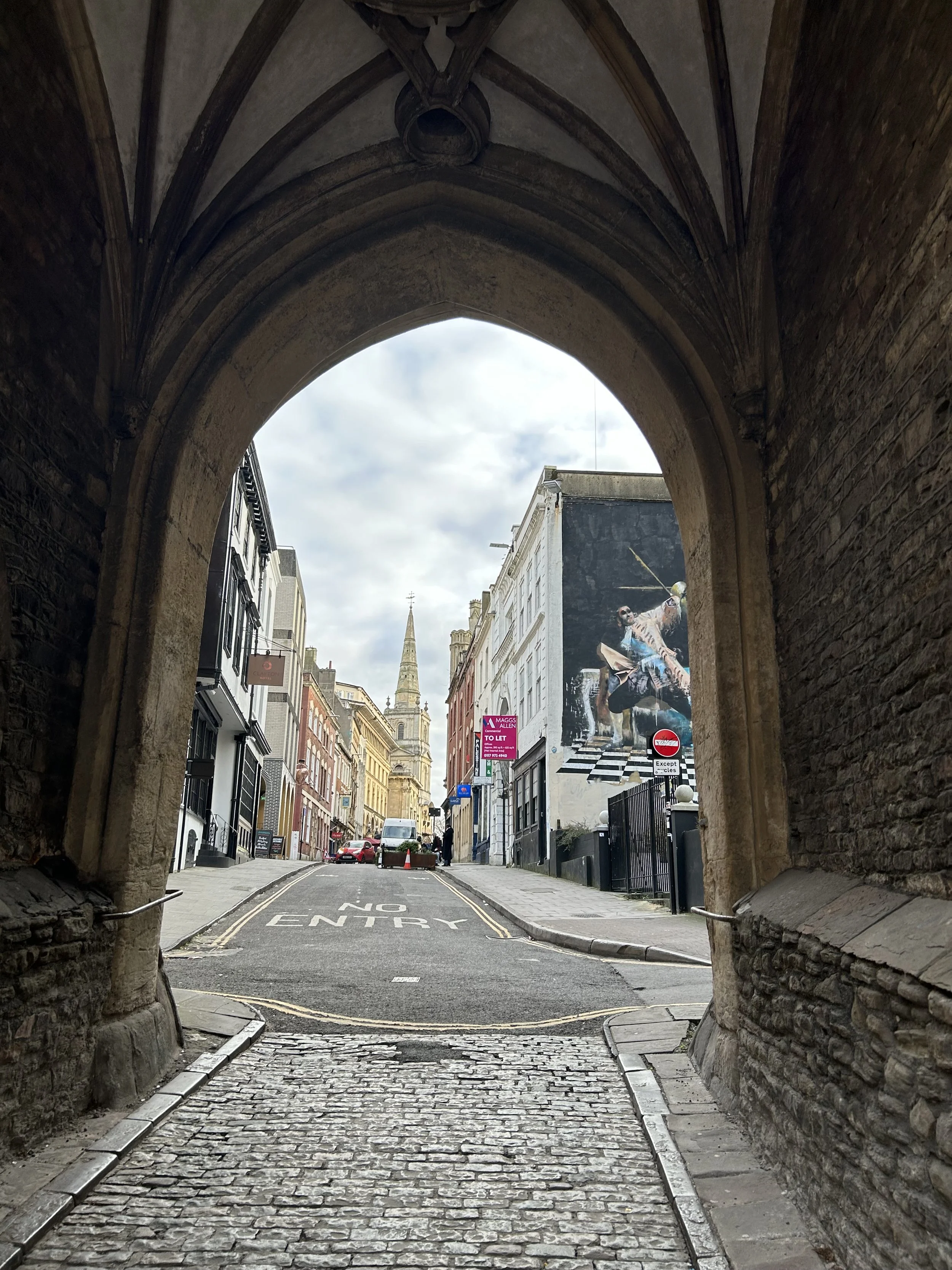 View of a cobblestone street through a stone archway, with colorful buildings, parked cars, and a church steeple in the distance. A large mural of a man sitting on a chair, playing guitar, is on the right side of the street.