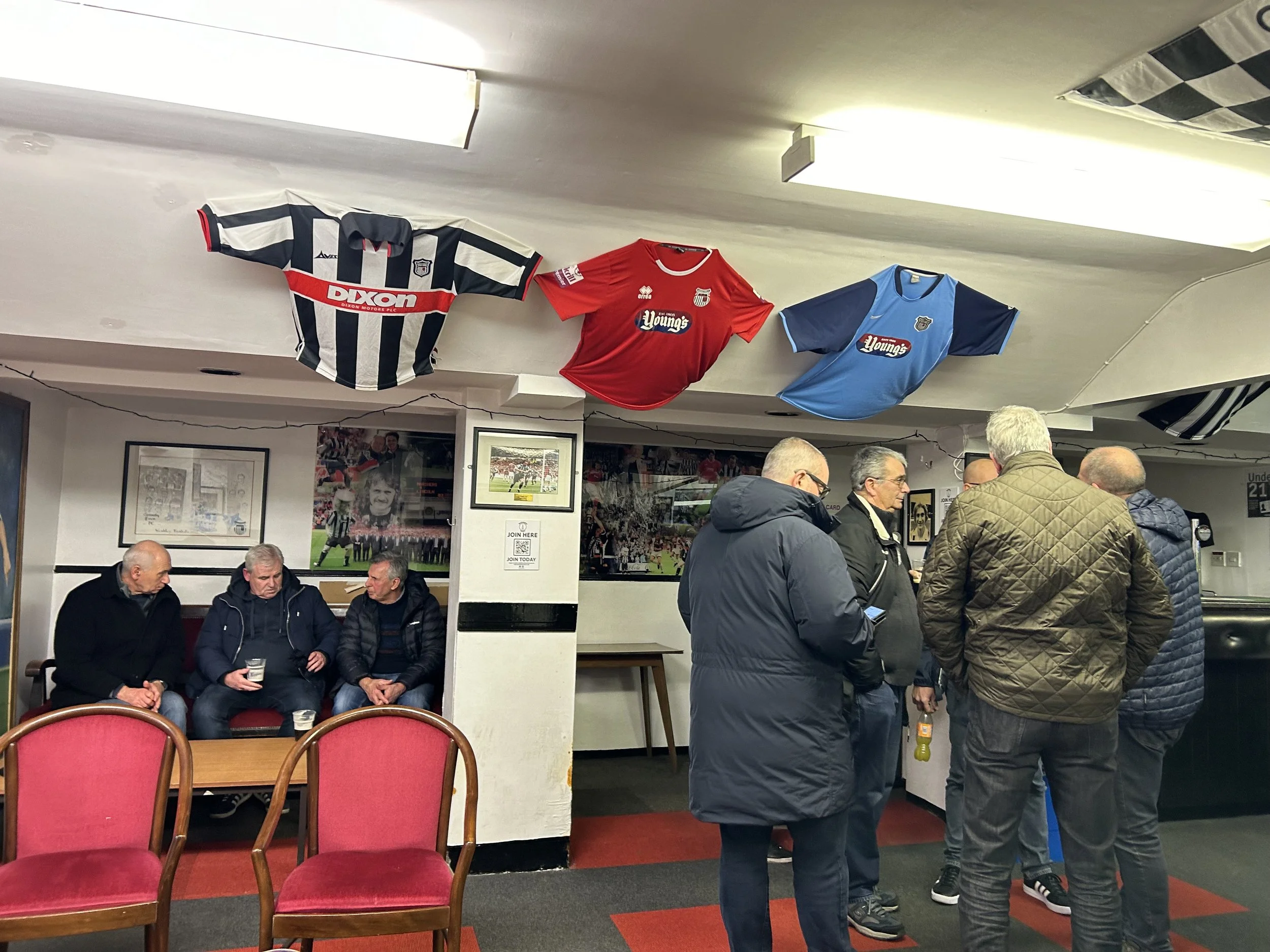 People socializing in a sports bar with framed sports photos on the wall and three sports jerseys hanging from the ceiling.