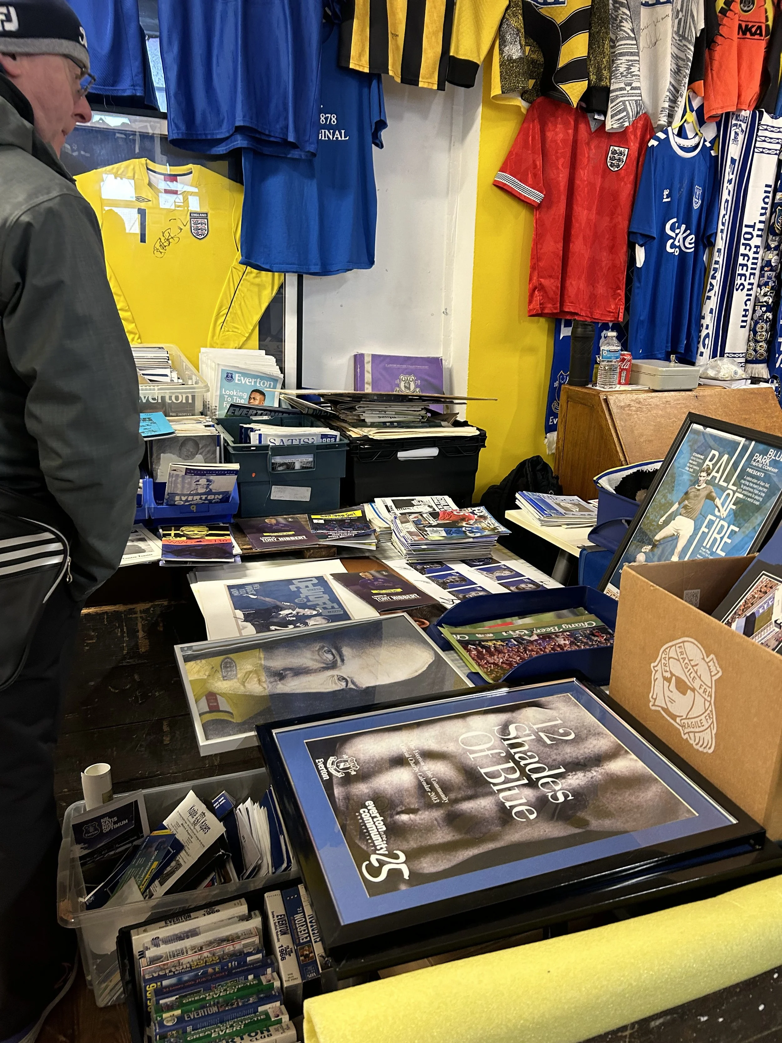 A memorabilia and merchandise table at a sports event or store, featuring framed photos of soccer players such as David Beckham, various soccer jerseys hanging on the wall including England and other teams, and stacks of magazines, brochures, and col