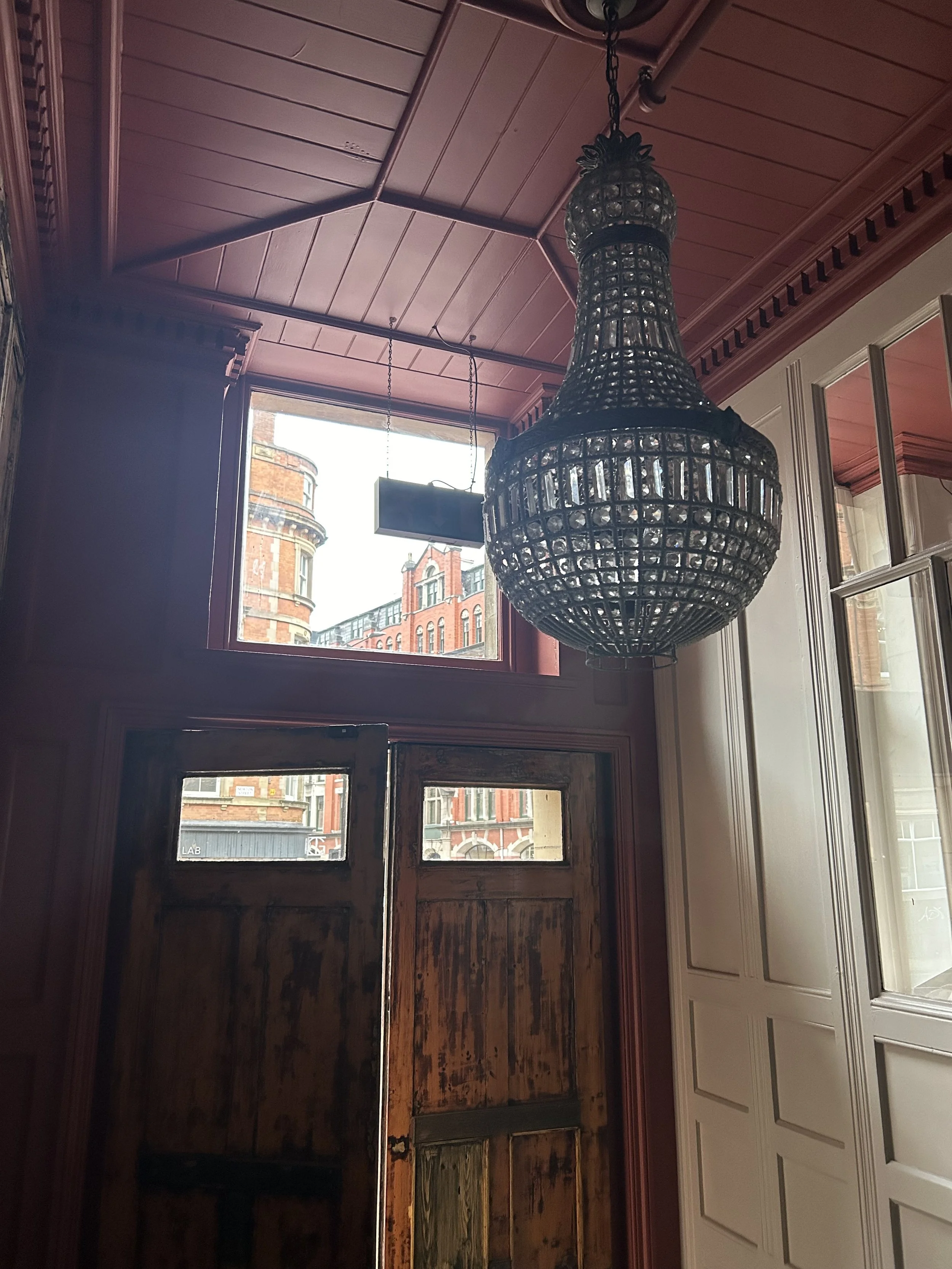Interior view of a room with a large ornate chandelier hanging from a pink wooden ceiling, a wooden door with peeling paint, and a window showing buildings outside.