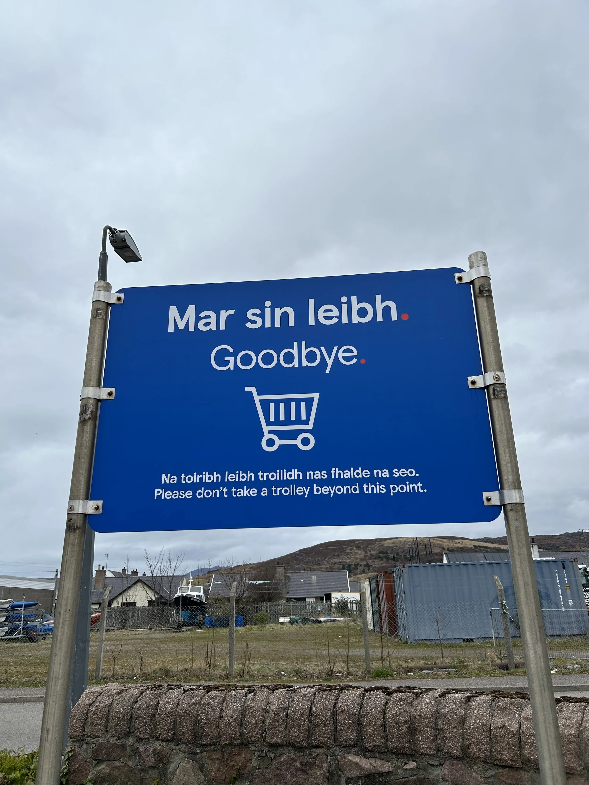Blue sign with white text and a shopping cart icon. The text reads, 'Mar sin leibh. Goodbye.' Below, it says, 'Na toirbh leibh troilidh nas fhaide na seo. Please don't take a trolley beyond this point.' in Gaelic and English. The background shows a g