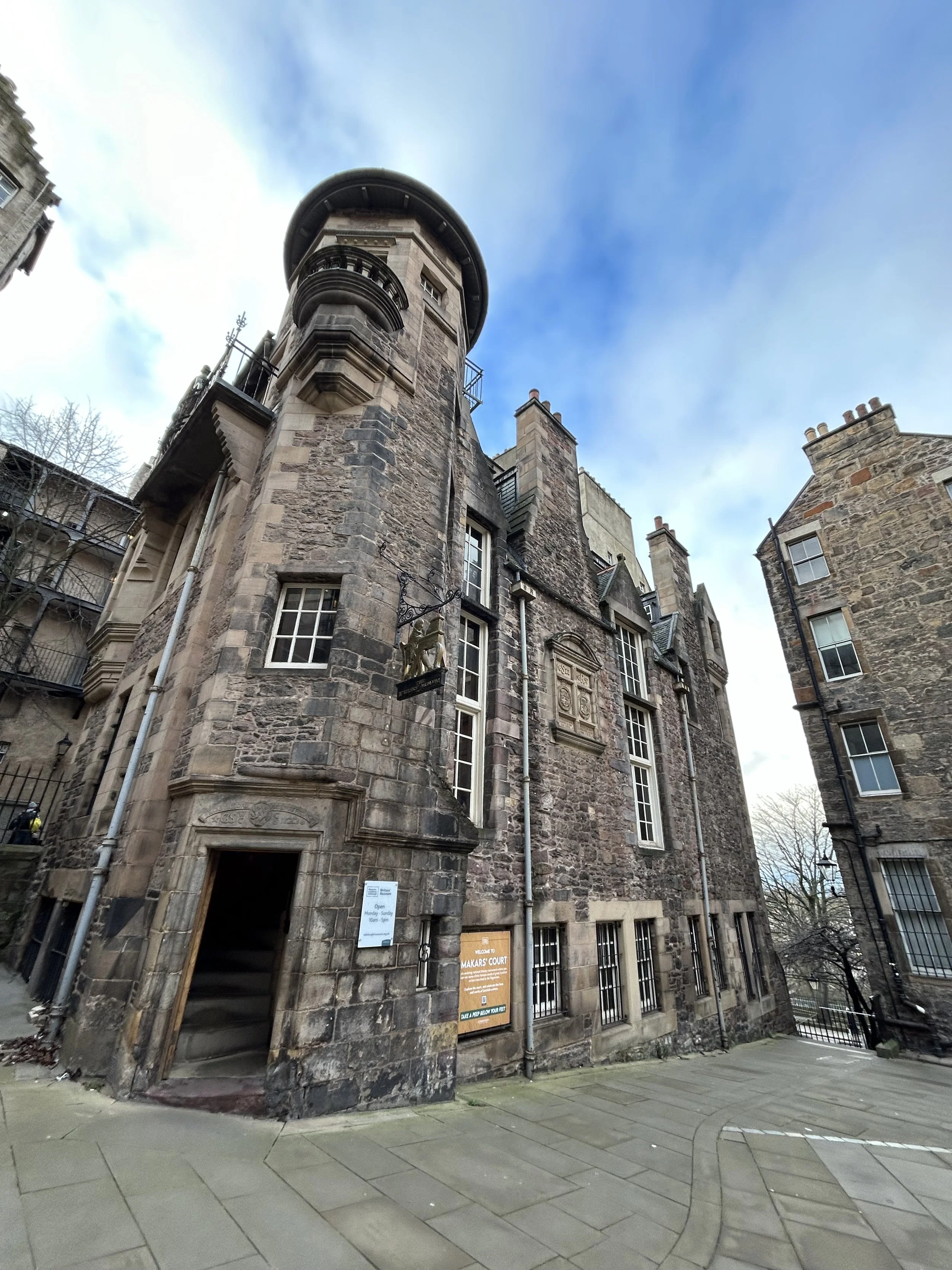 Historic stone building with a round tower, decorative carvings, and tall windows, located at the corner of a street with a paved sidewalk and neighboring buildings under a partly cloudy sky.