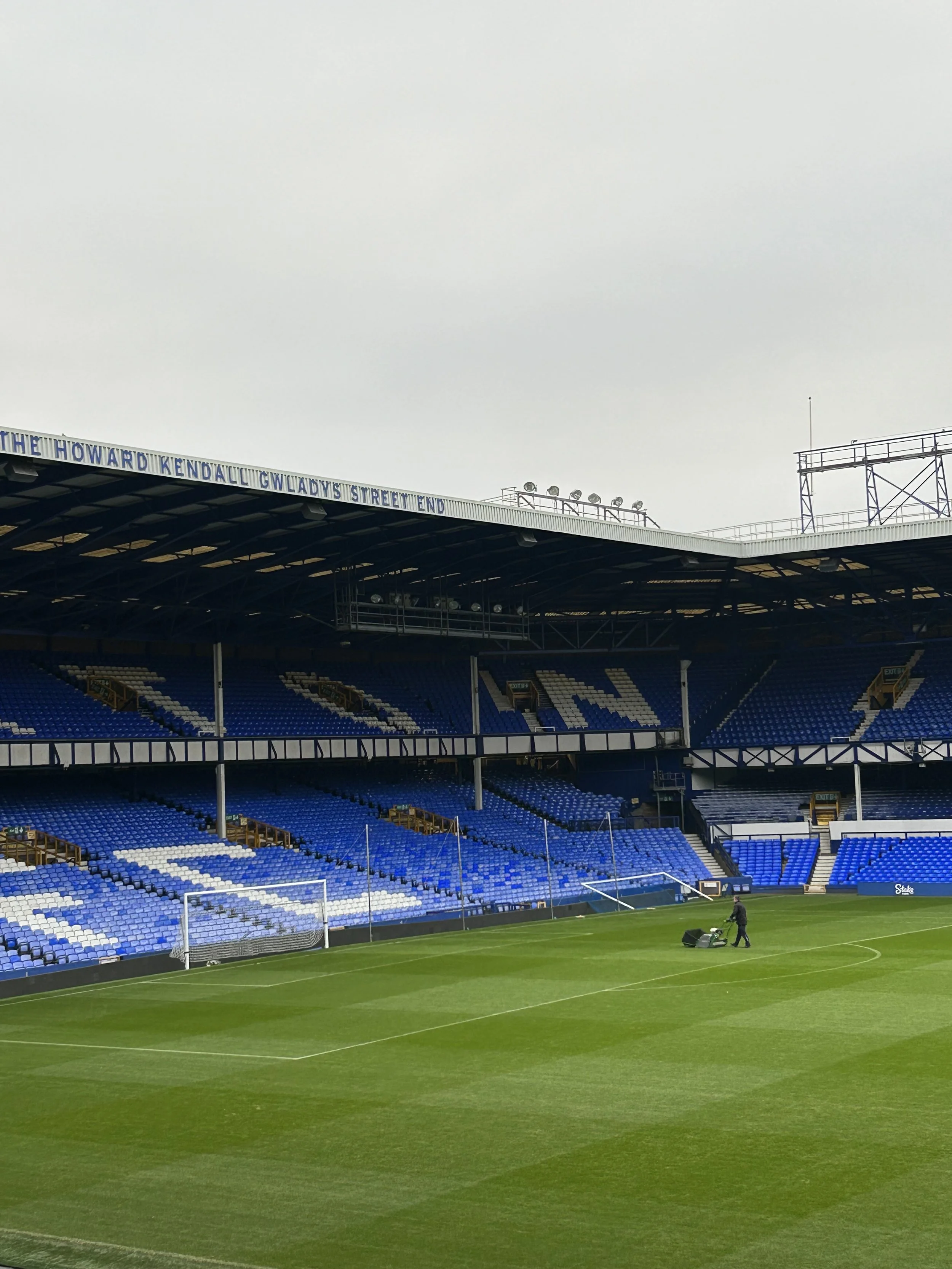Empty football stadium with green field, blue and white seats, and overcast sky.
