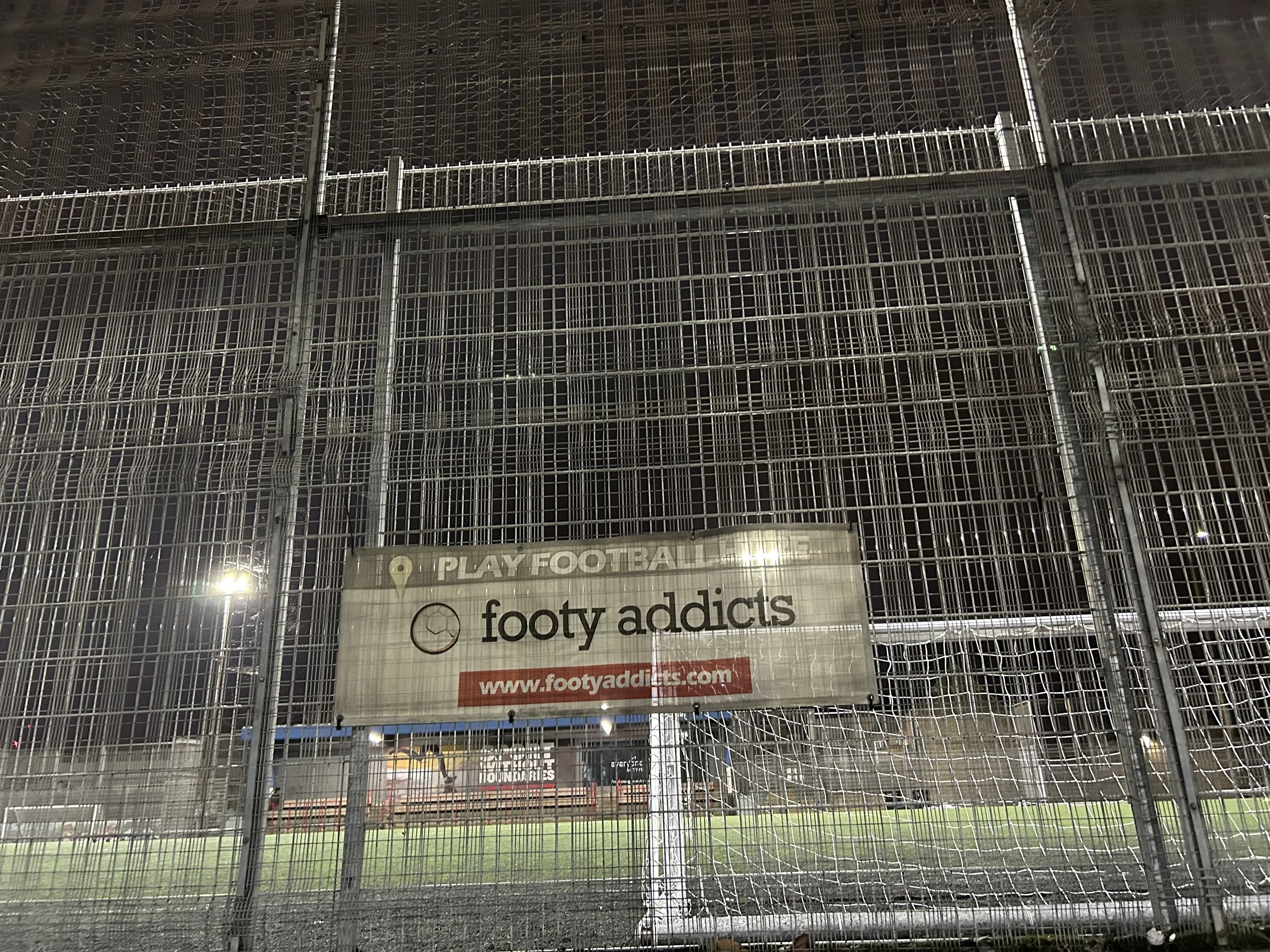 A metal fence with a sign that reads 'footy addicts' and a website URL, with a football field visible behind the fence.