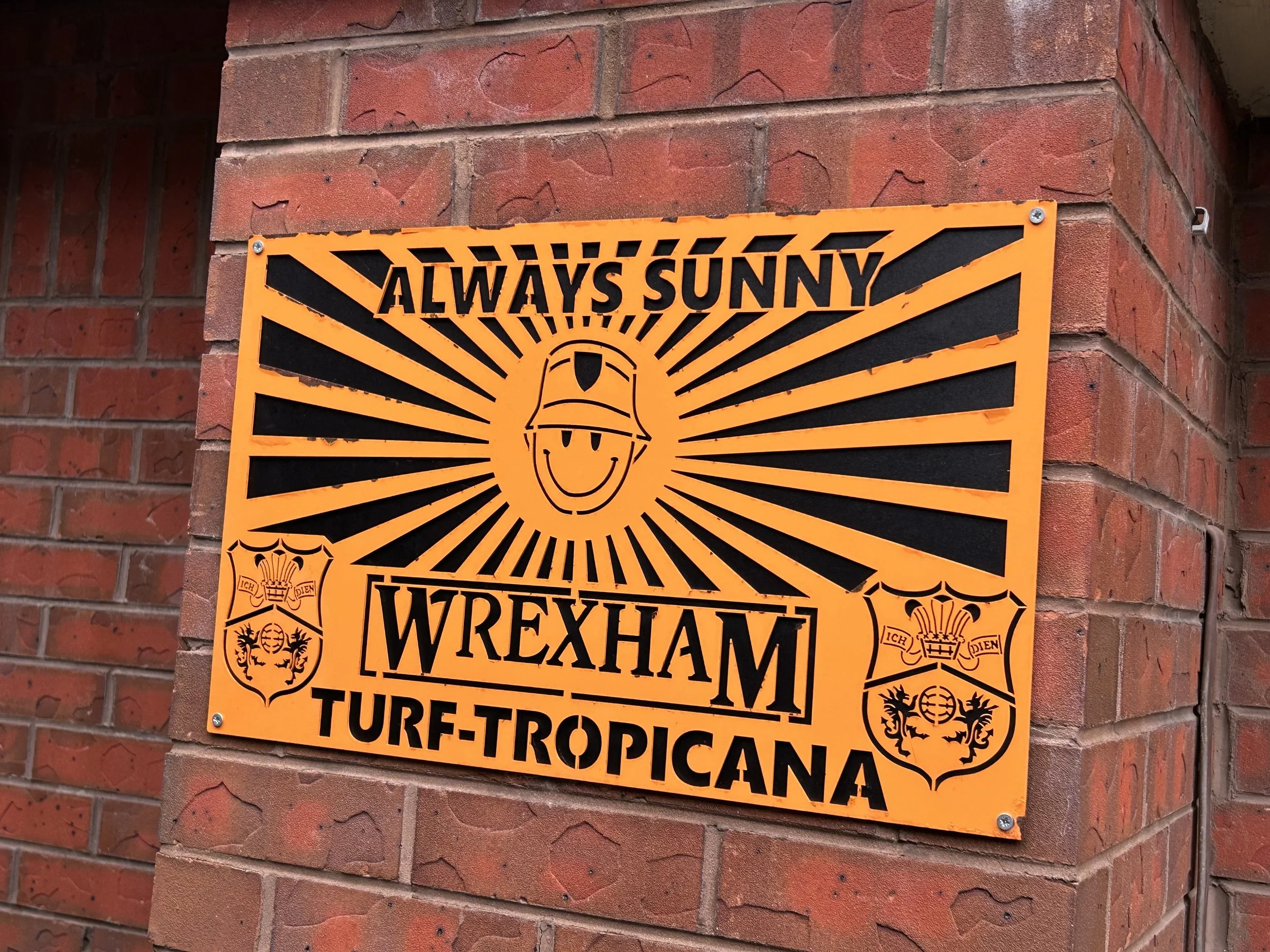 A decorative orange and black sign affixed to a brick wall, featuring a smiling cartoon face wearing a construction helmet, with radiant lines emanating from it. The sign reads "ALWAYS SUNNY WREXHAM TURF-TROPICANA" and includes crest-like emblems on 