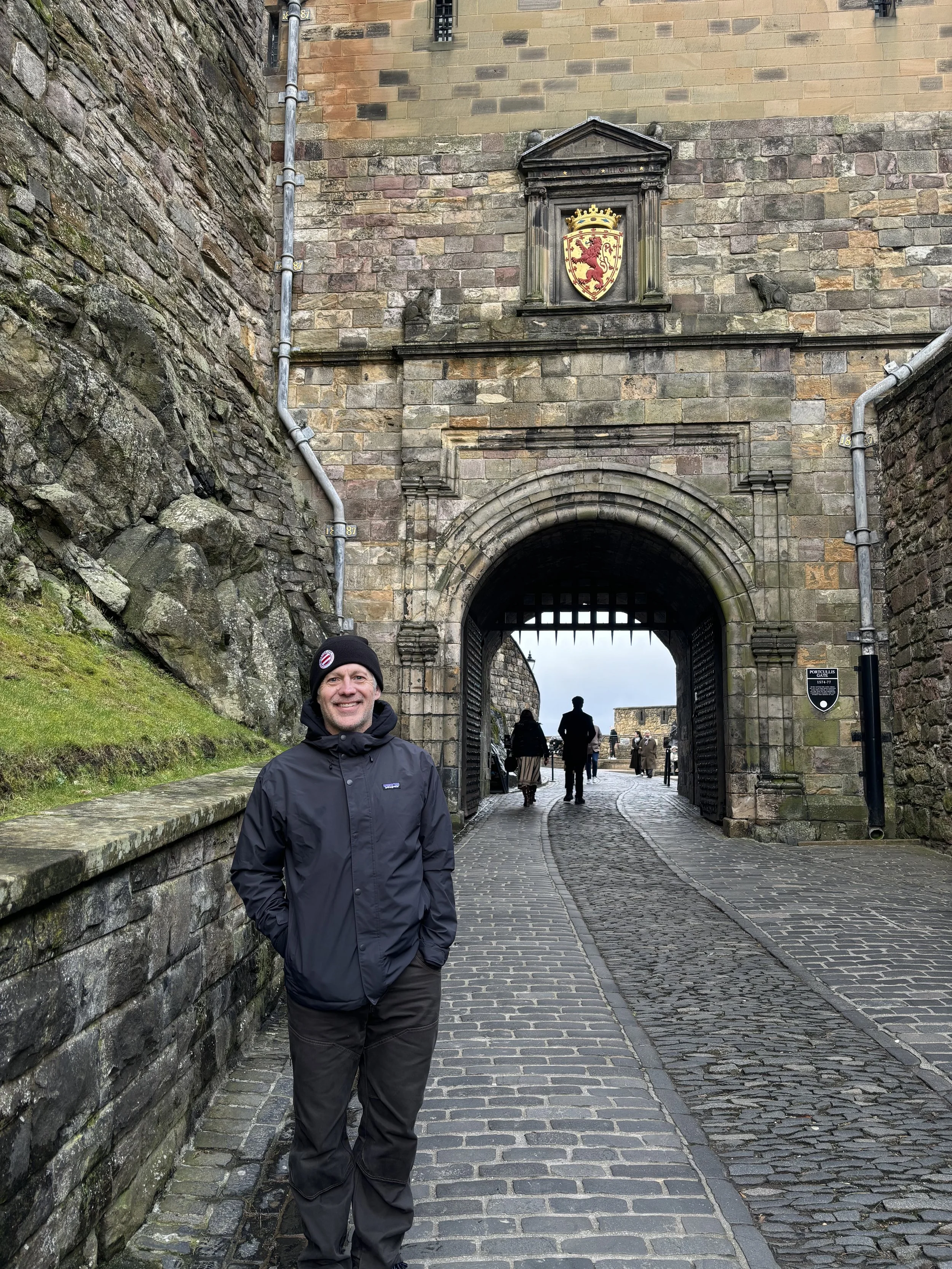 A man in a black jacket and beanie standing outside the entrance of a historic stone castle gate with a flag and crest above it, holding his hands in his pockets and smiling.