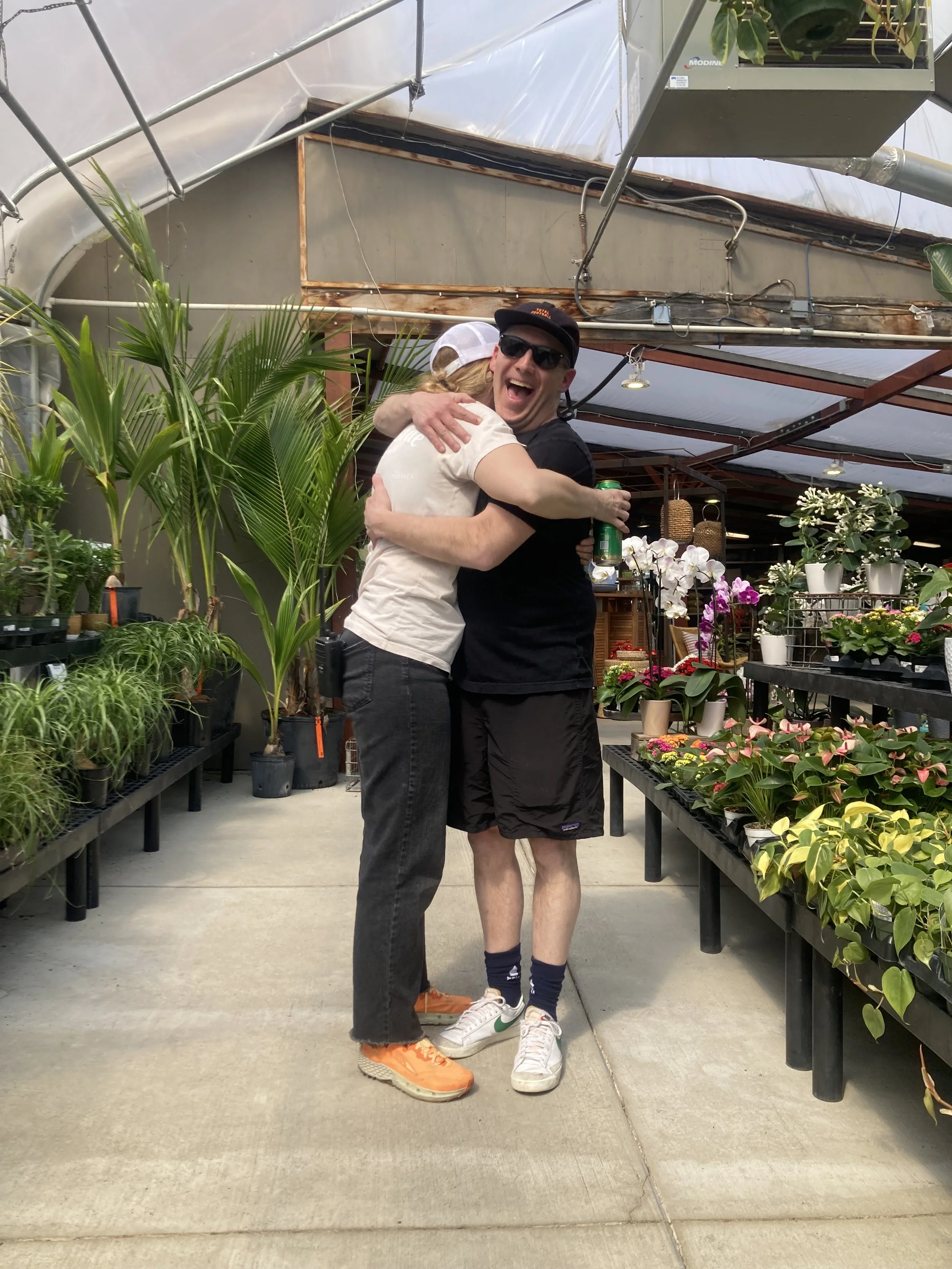 Two people hugging inside a greenhouse surrounded by potted plants and flowers, one wearing a white baseball cap and the other wearing sunglasses and a black cap.