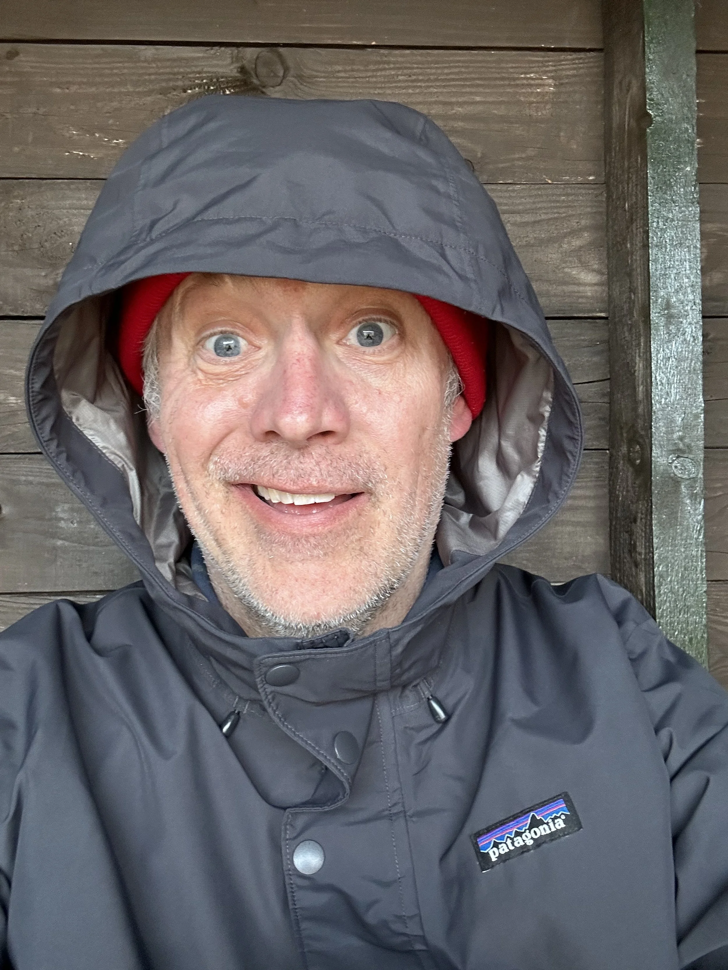 A man with gray hair, wearing a red beanie and a black Patagonia jacket with a hood, smiling in front of a wooden background.