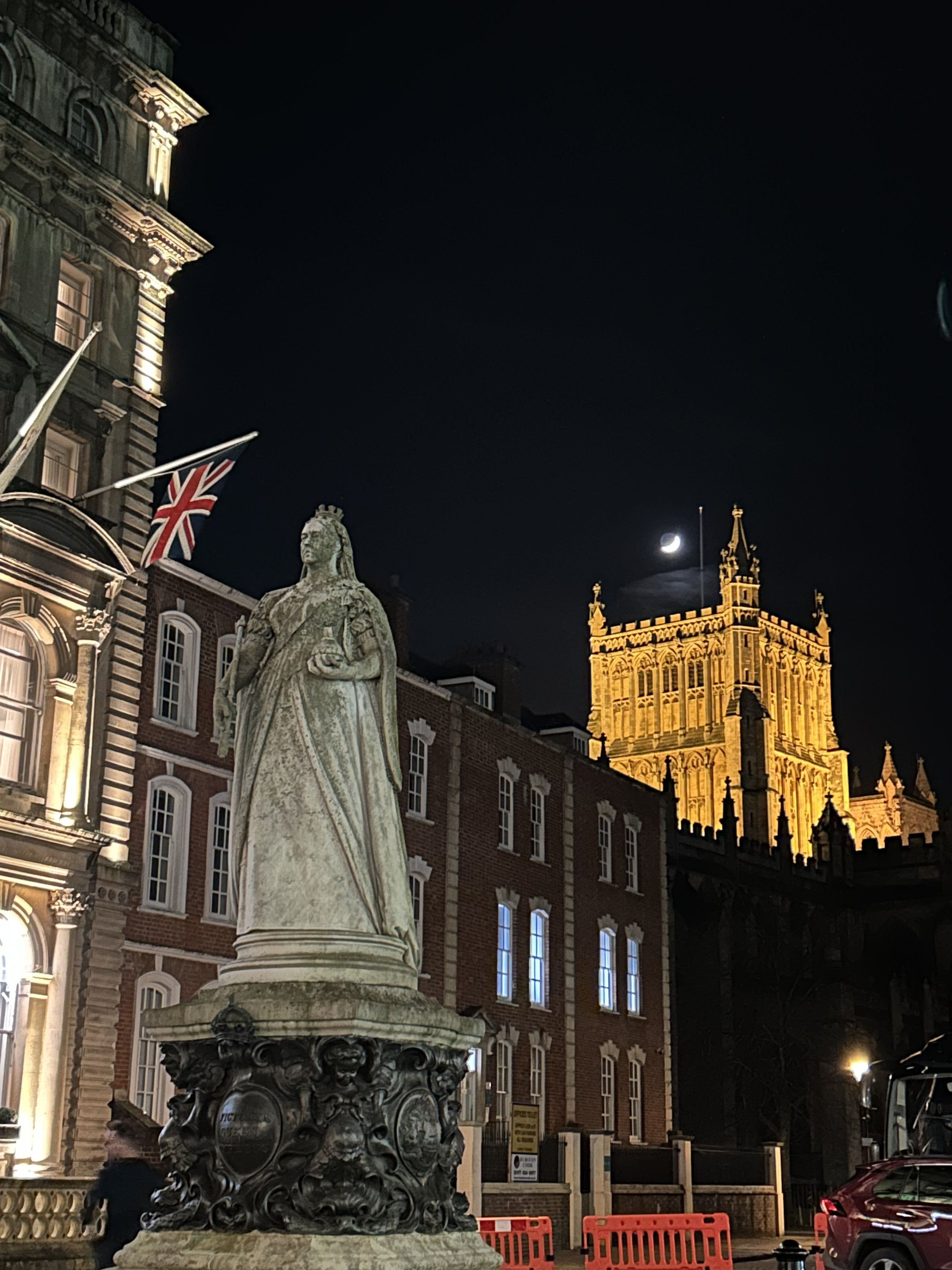 Nighttime scene of historic buildings in London, featuring a statue of Queen Victoria in the foreground, the illuminated Victoria Tower of the Palace of Westminster in the background, a Union Jack flag, and a crescent moon in the dark sky.