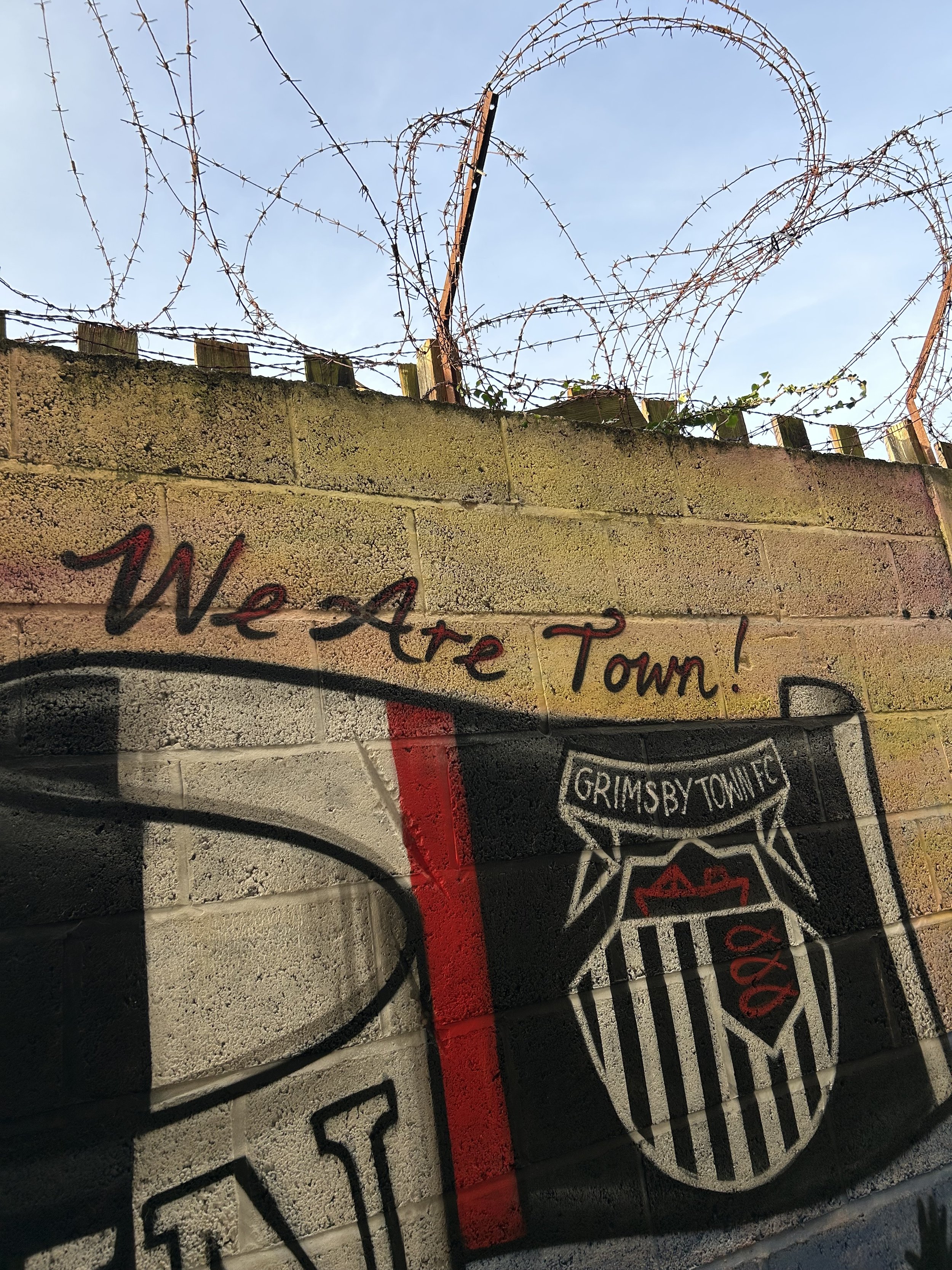 Graffiti on a brick wall that says "We Are Town!" along with a shield logo that reads "Grimsby Town FC" and features a red and white design. Barbed wire is visible above the wall, with a clear sky in the background.