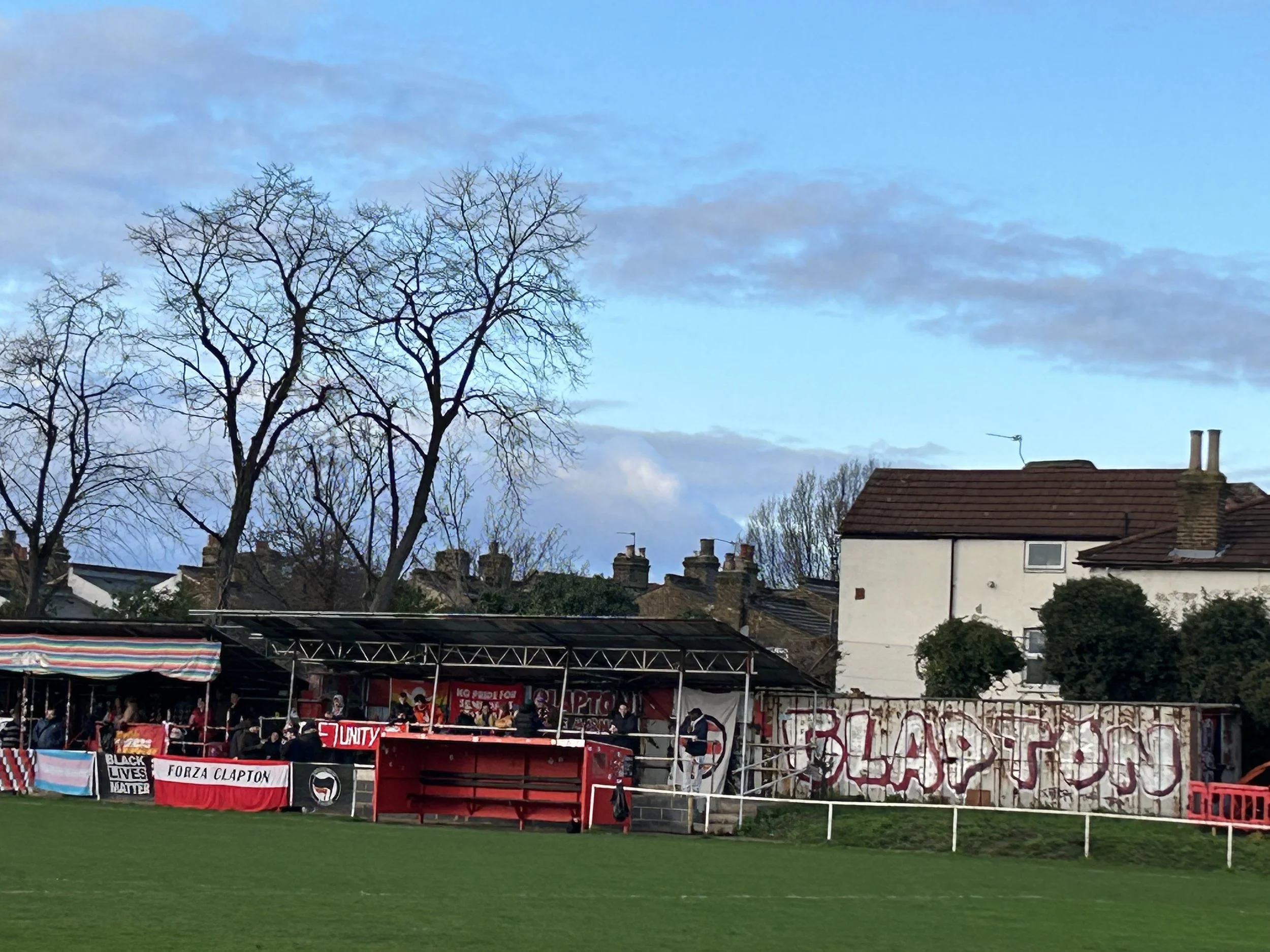 A small football stadium with a red covered seating area, banners with messages including 'Black Lives Matter' and 'Forza Clapton,' and graffiti on a wall that reads 'Clapton.'