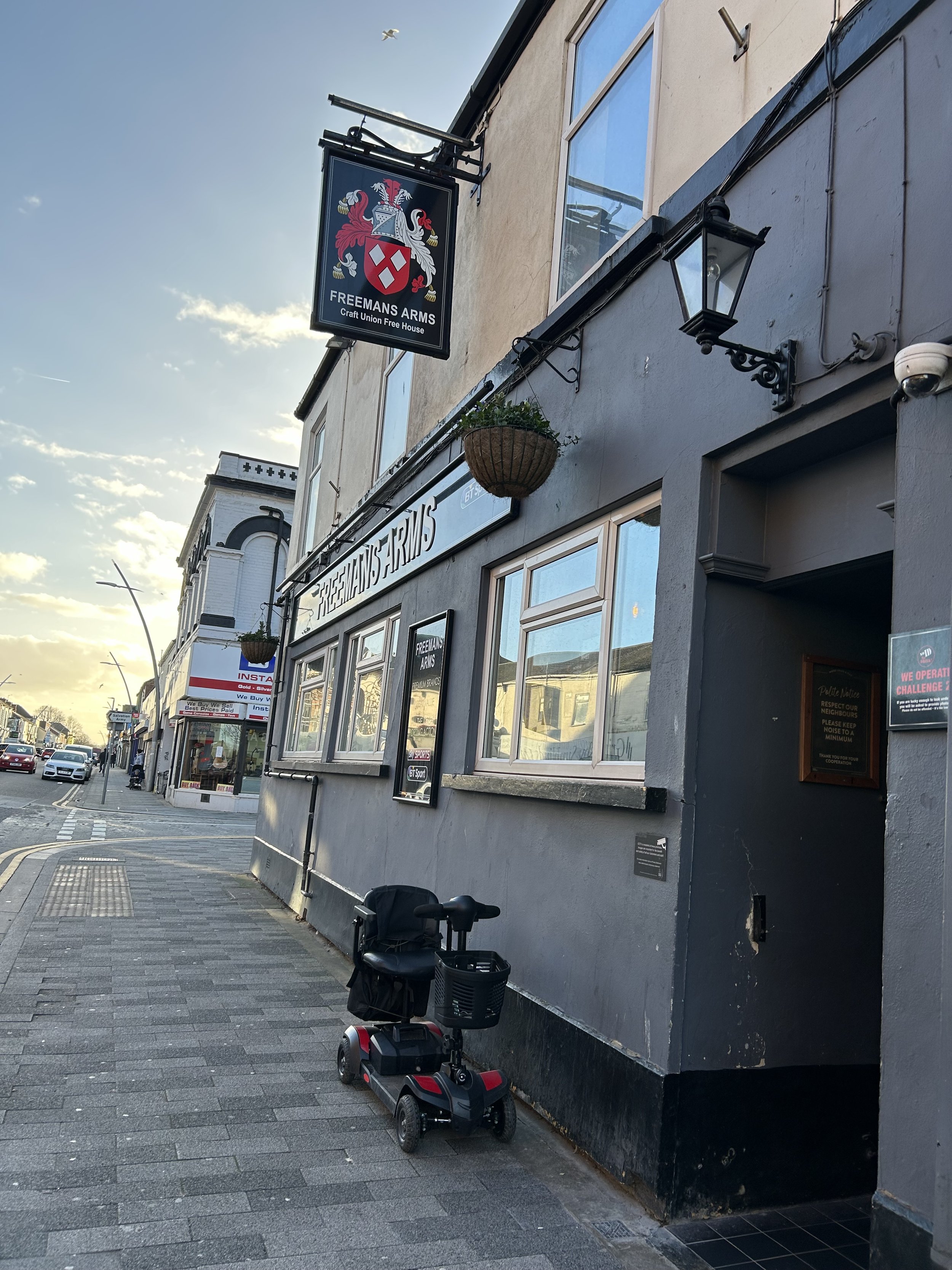 Street view showing a gray building with a sign for Freemans Arms, a pub, with a scooter parked outside on the sidewalk. The sky is partly cloudy with sunlight.