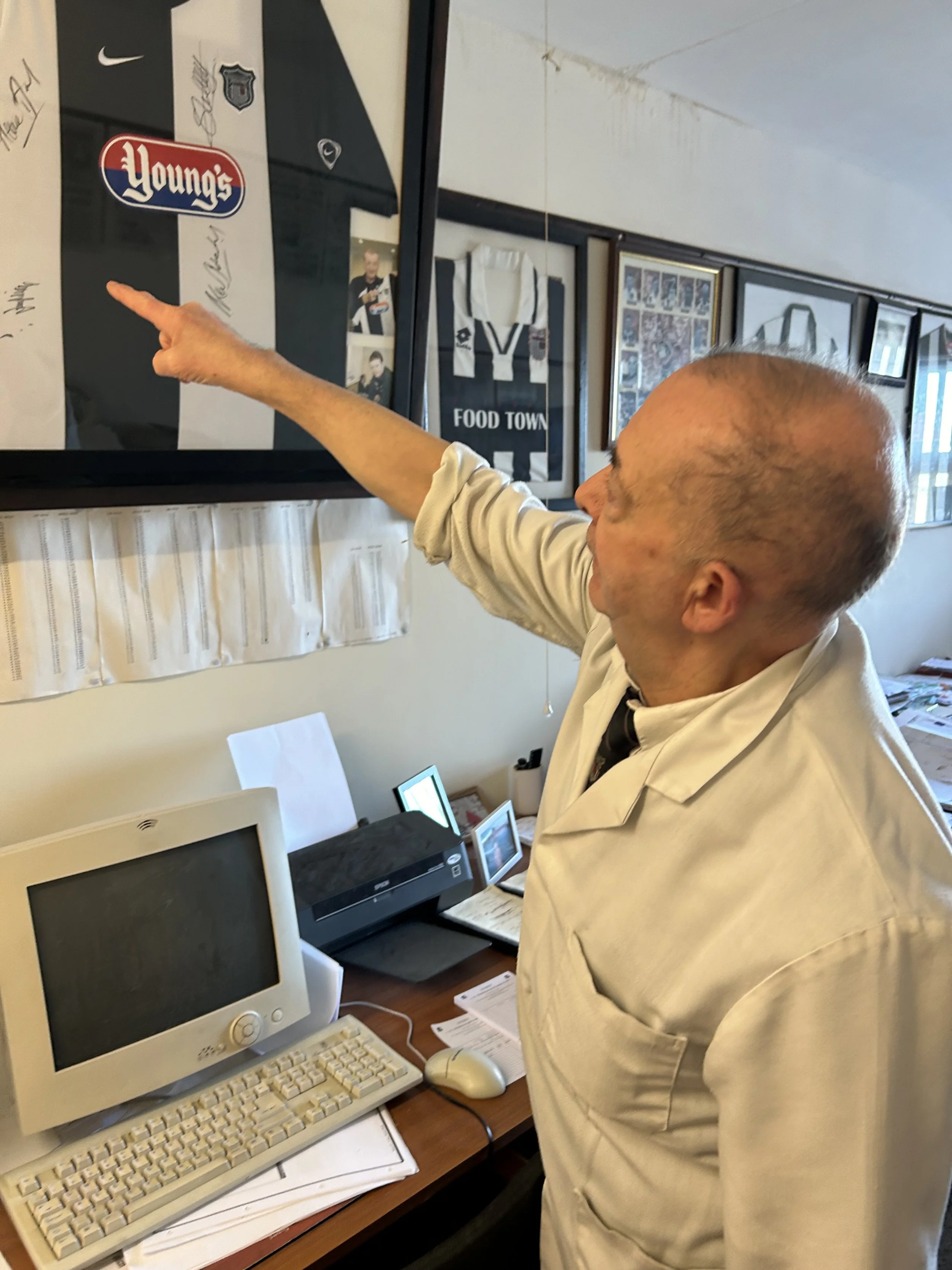An elderly man in a white lab coat pointing at a framed item on the wall in an office or memorabilia room.