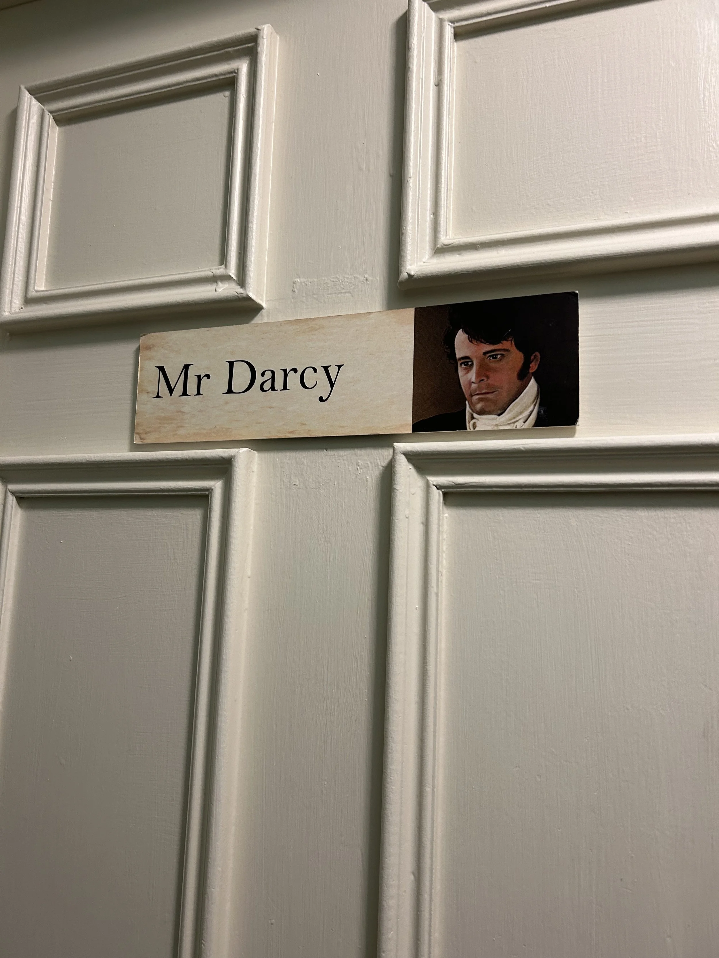 Close-up of a white door with a nameplate reading 'Mr Darcy' and a picture of a man with dark, curly hair and period clothing.