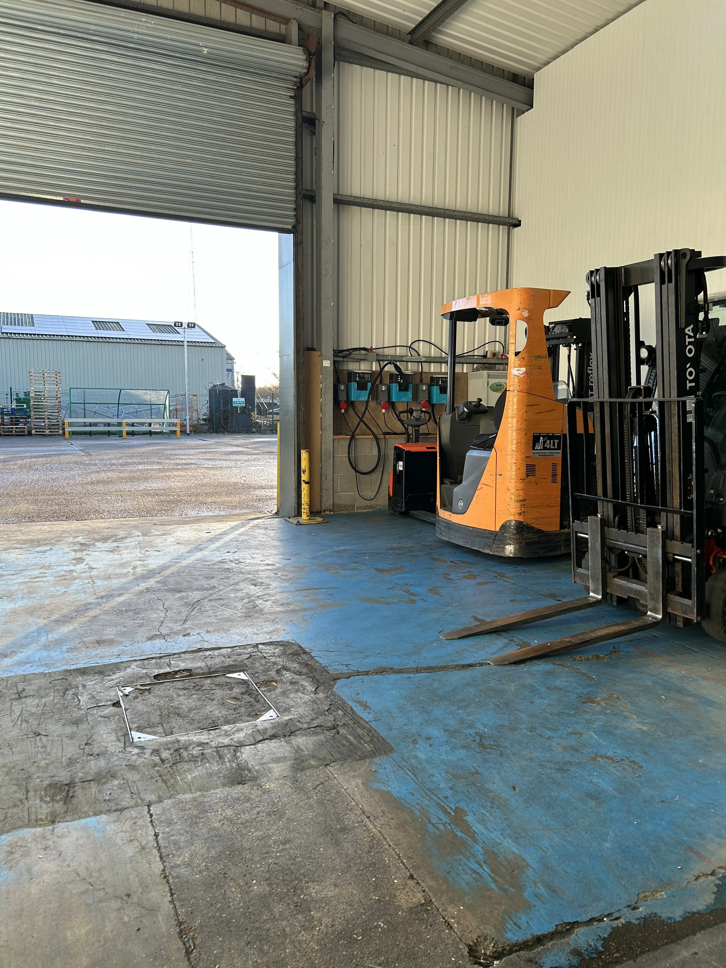 Inside of a warehouse with a forklift parked near an open roller shutter door, allowing natural light to illuminate the space. There are electrical panels on the wall and a large empty floor area with visible cracks, paint, and a square hatch in the 