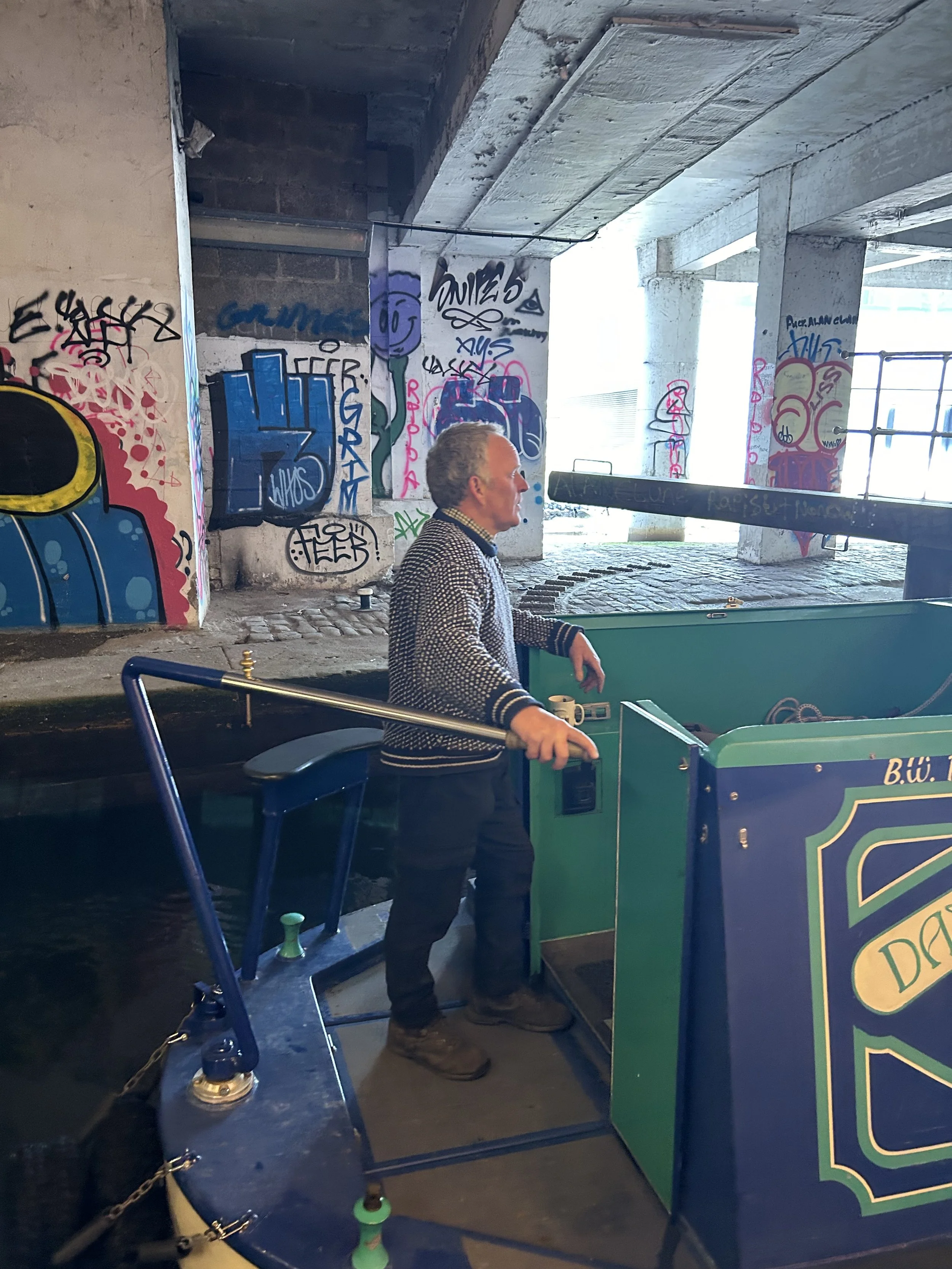 Man standing on a boat, operating a pinball machine in an urban setting with graffiti-covered walls and a canal nearby.