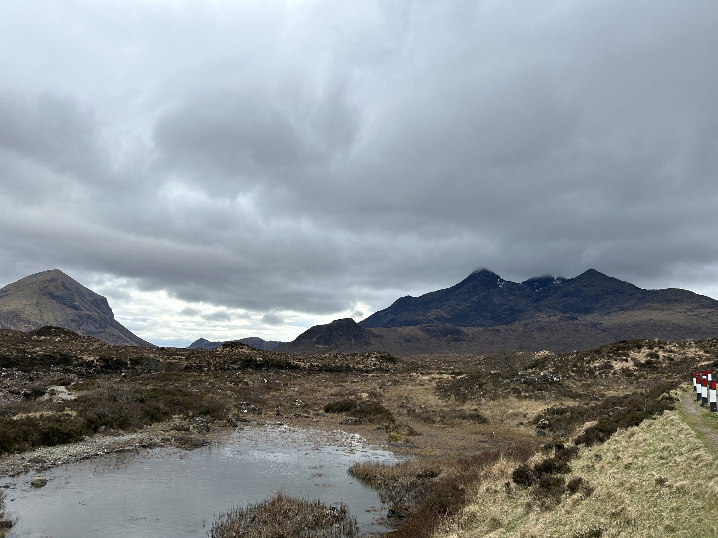 A mountainous landscape on a cloudy day with a small pond in the foreground and a grassy path with stone barriers on the right.