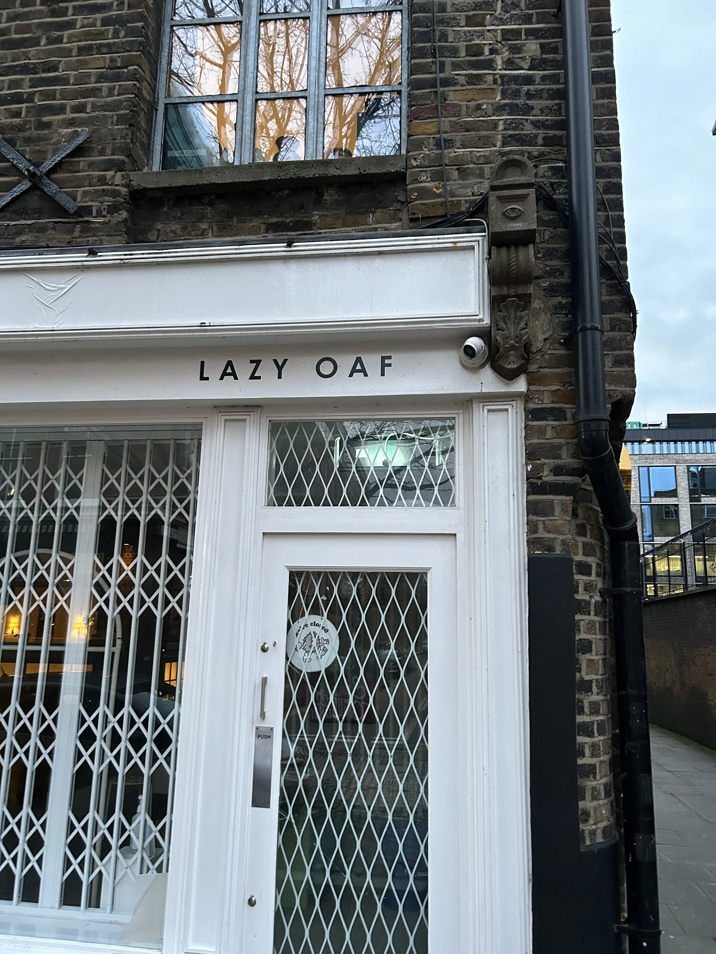 Close-up of a building entrance with a white sign reading "LAZY OAF" in black letters above a glass door secured with a metal grille, and a security camera to the right of the sign.