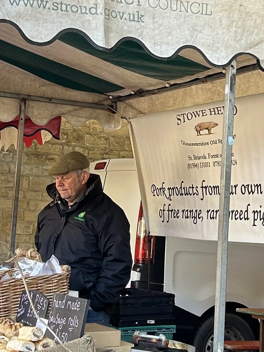 A man wearing a flat cap and black jacket standing behind a market stall selling pork pies and sausage rolls at an outdoor market. There are signs on the stall indicating the products and prices, and a large banner advertising pork products from a lo